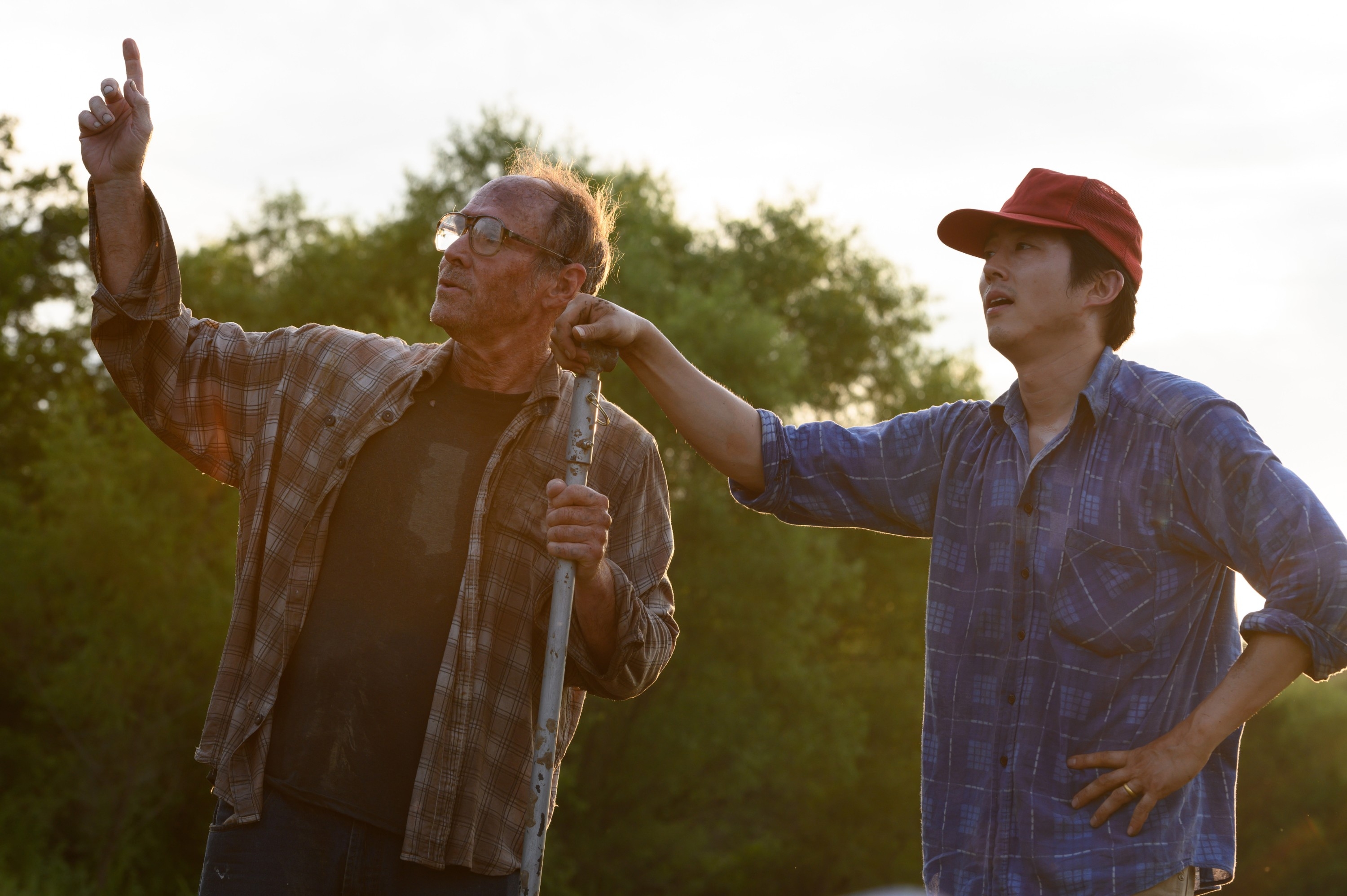Two men outdoors, one in a plaid shirt and cap pointing to the sky, the other observing with a thoughtful pose, set against a natural background