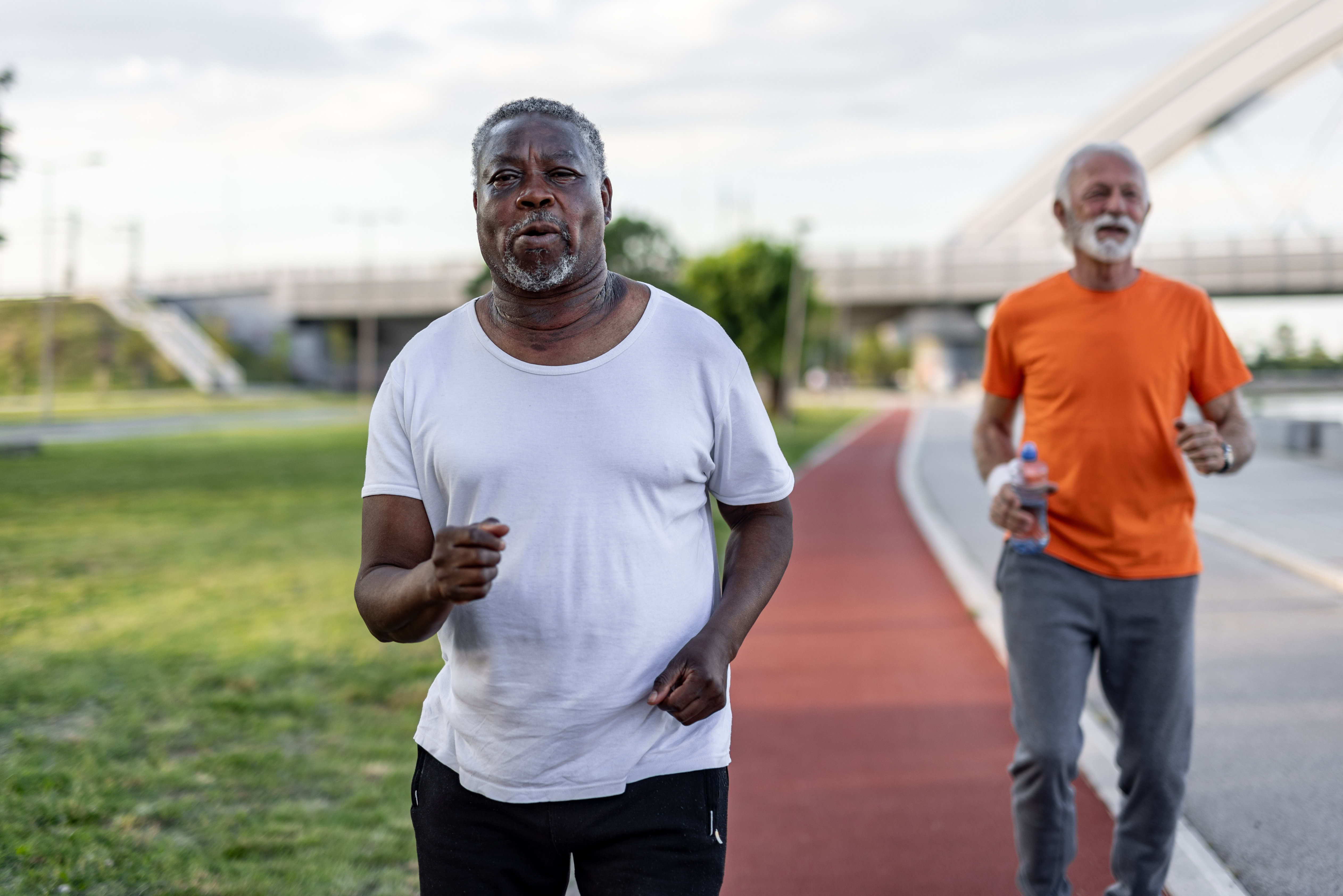 To menn som jogger på et spor, den ene i en hvit skjorte og den andre i en oransje skjorte, utendørs på en solskinnsdag
