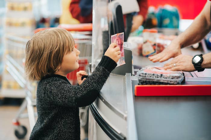 Child handing cash to a cashier at a grocery checkout. A plastic container of berries is on the counter
