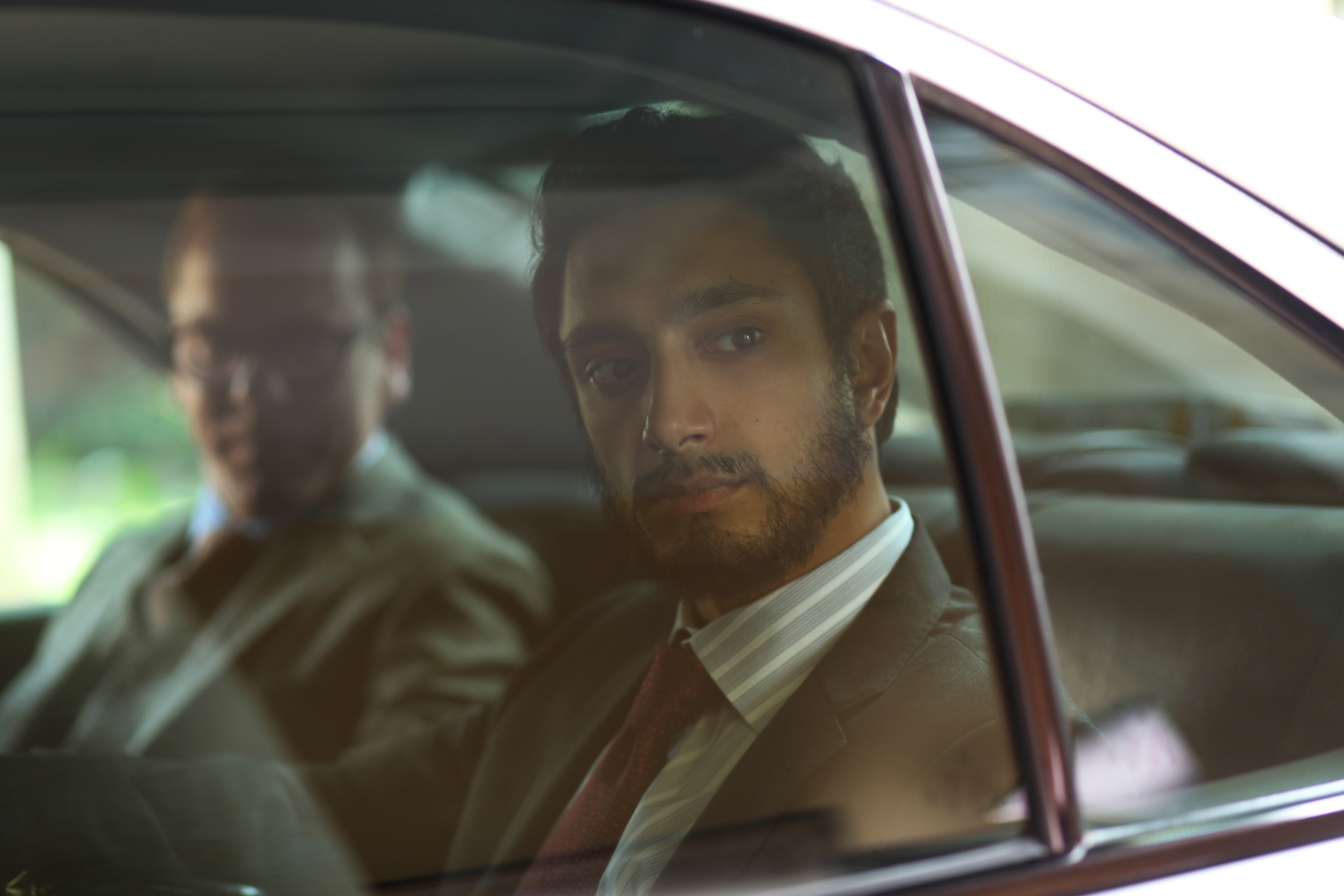 Two men sit in the backseat of a car, one looking contemplative out the window. The scene suggests a storyline or character moment from a movie or TV show