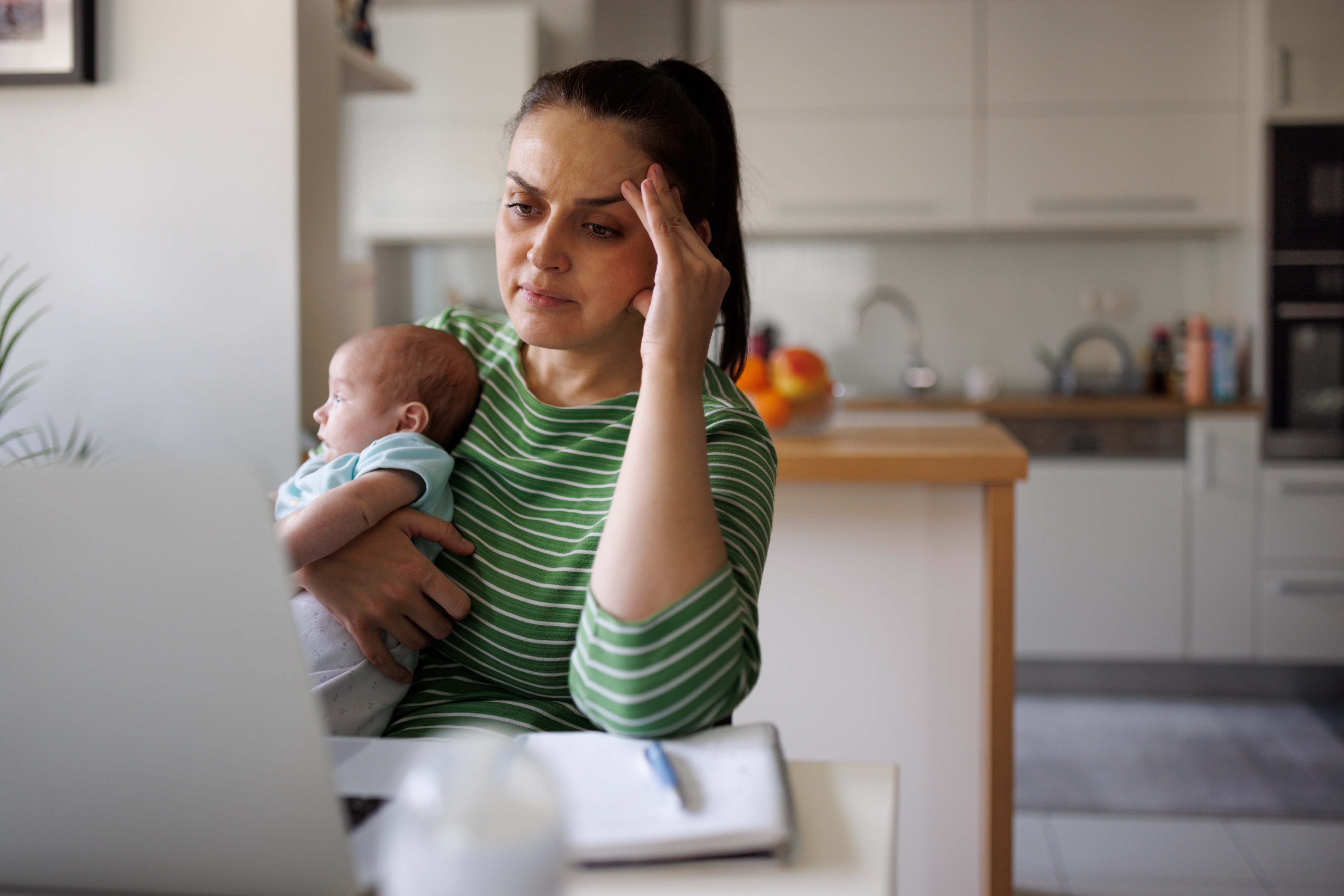 Woman cradling a baby looks at a laptop screen with a concerned expression, seated at a kitchen table with a notebook and pen nearby