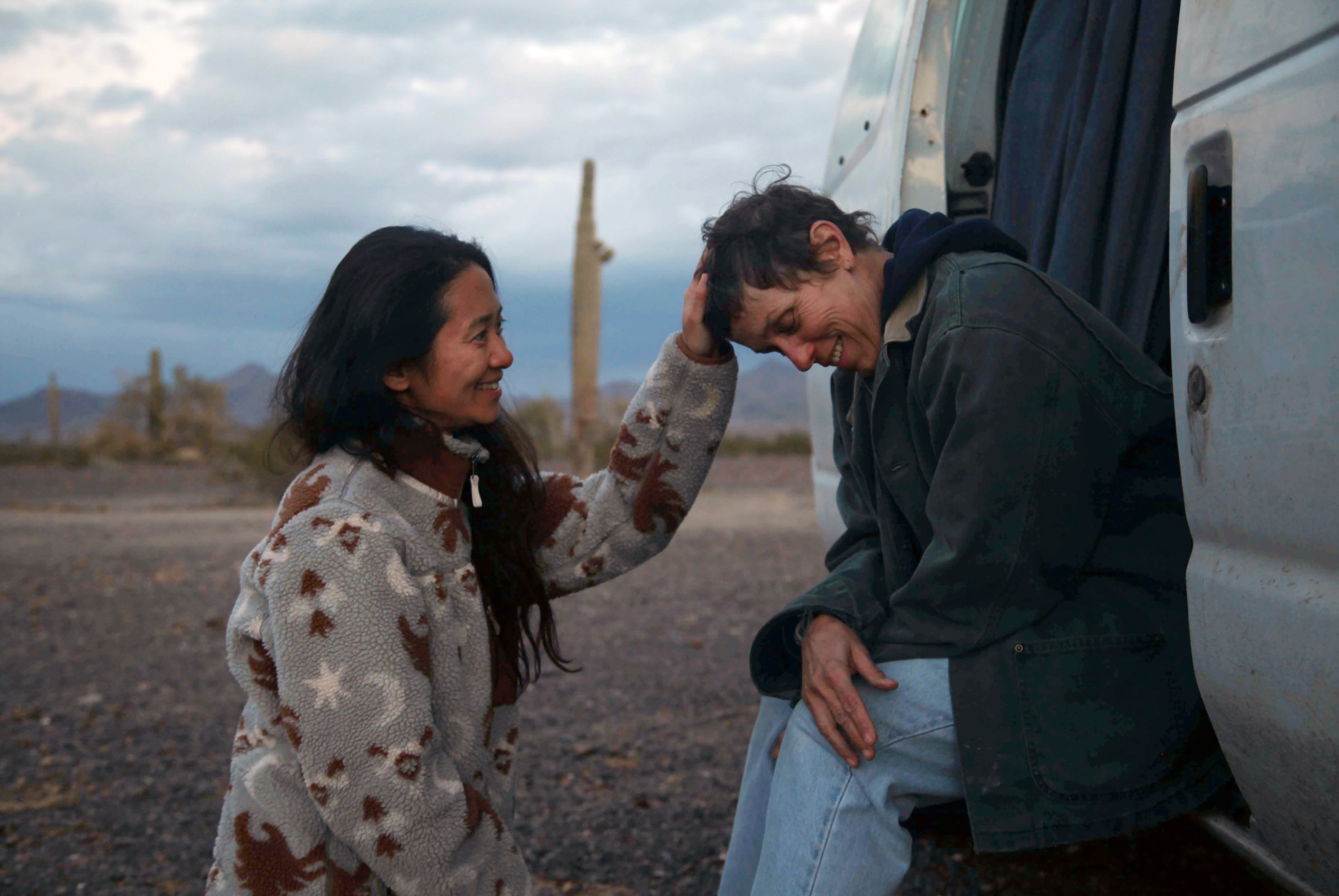 Two people share a warm moment beside a van in a desert setting, with one gently touching the other's head, creating a sense of companionship