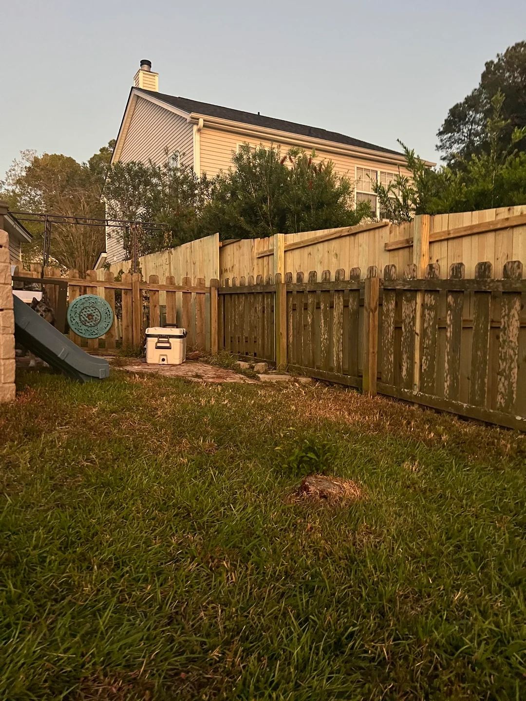 Backyard scene with a slide, cooler, and wooden fence. suburban house and trees in the background