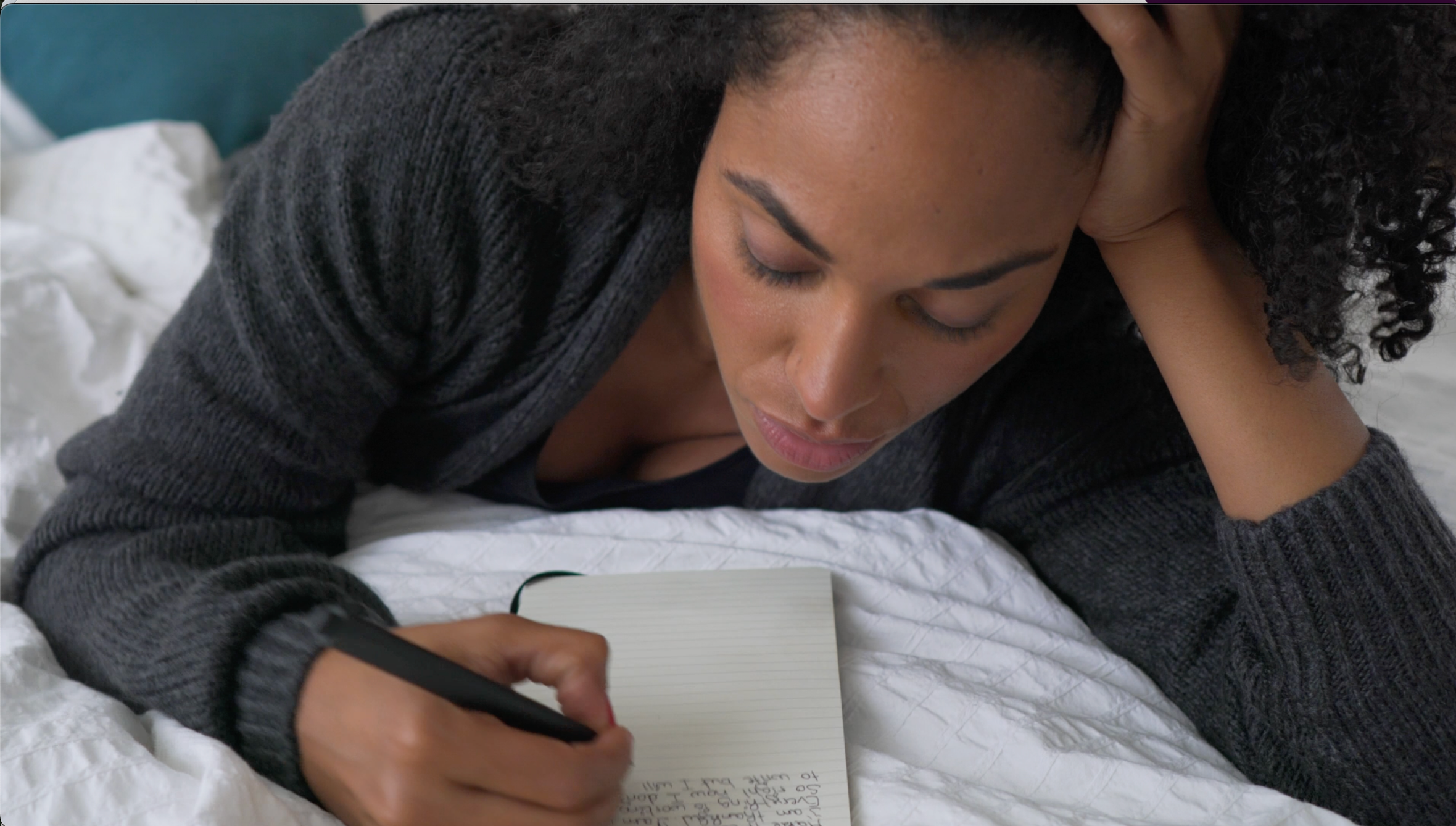 Person writing in a notebook while lying on a bed, wearing a sweater, looking thoughtful