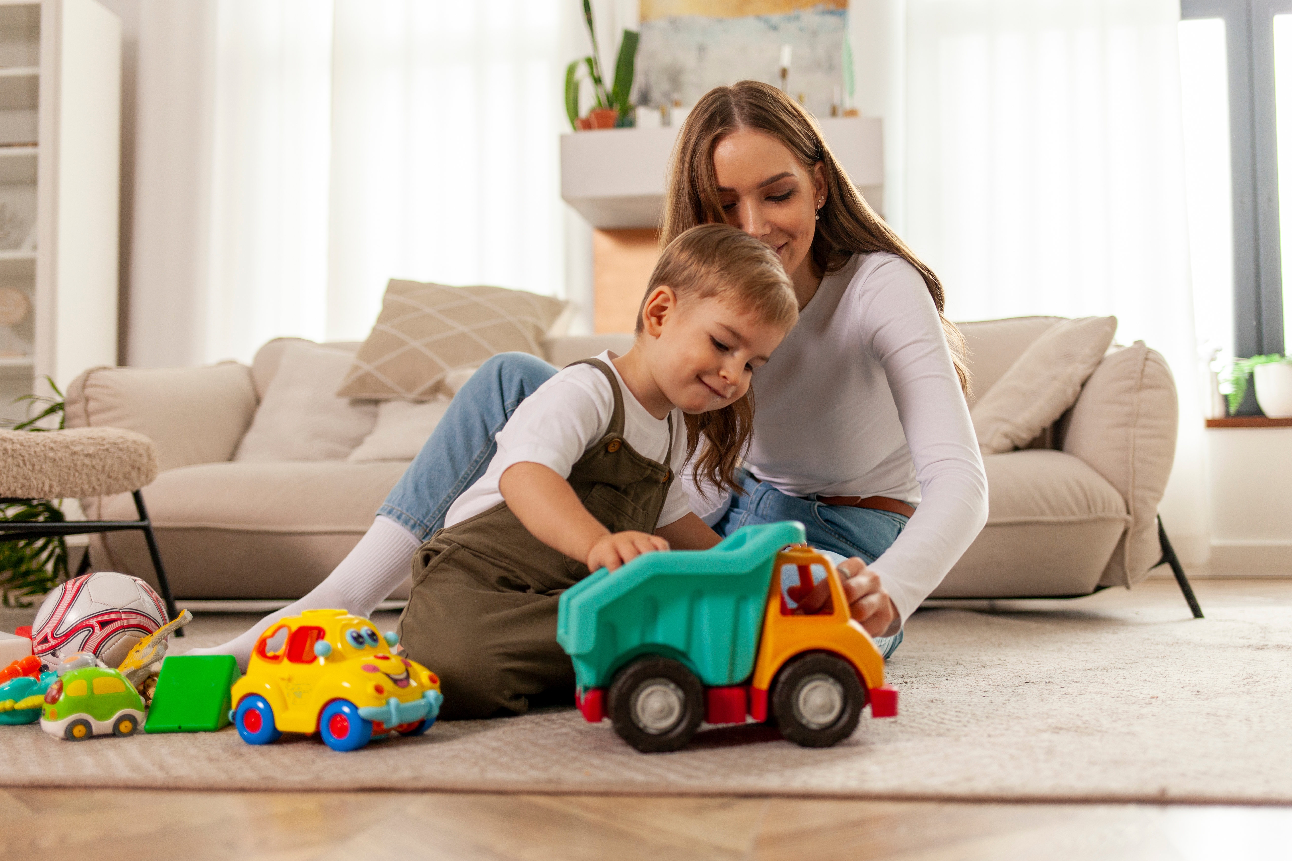 A woman and child sit on the floor, engaging in play with toy vehicles in a cozy living room setting