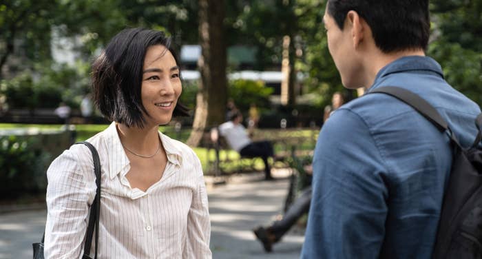 Two people are having a conversation in a park. One, with a short hairstyle, wears a casual shirt. The other has their back to the camera