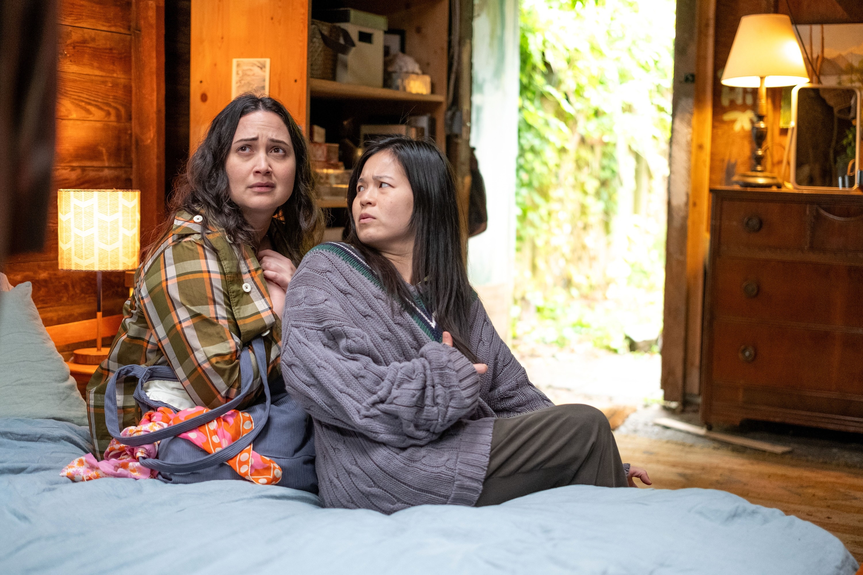 Two women sit on a bed in a cozy room, looking concerned. One wears a plaid jacket, and the other a gray sweater, suggesting a tense moment