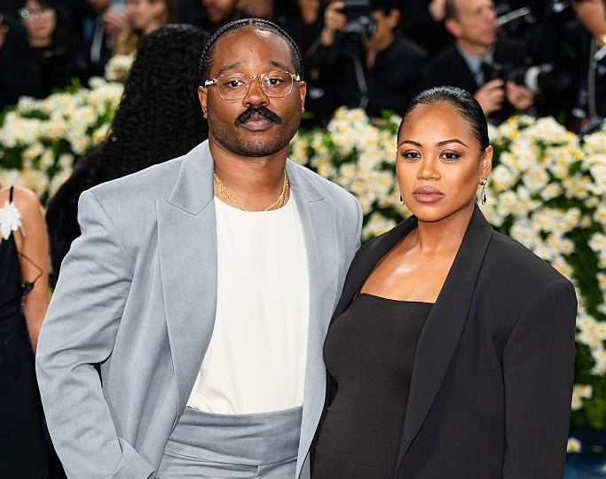 A couple poses at a red carpet event. The man wears a light suit, and the woman is in a black dress with a blazer
