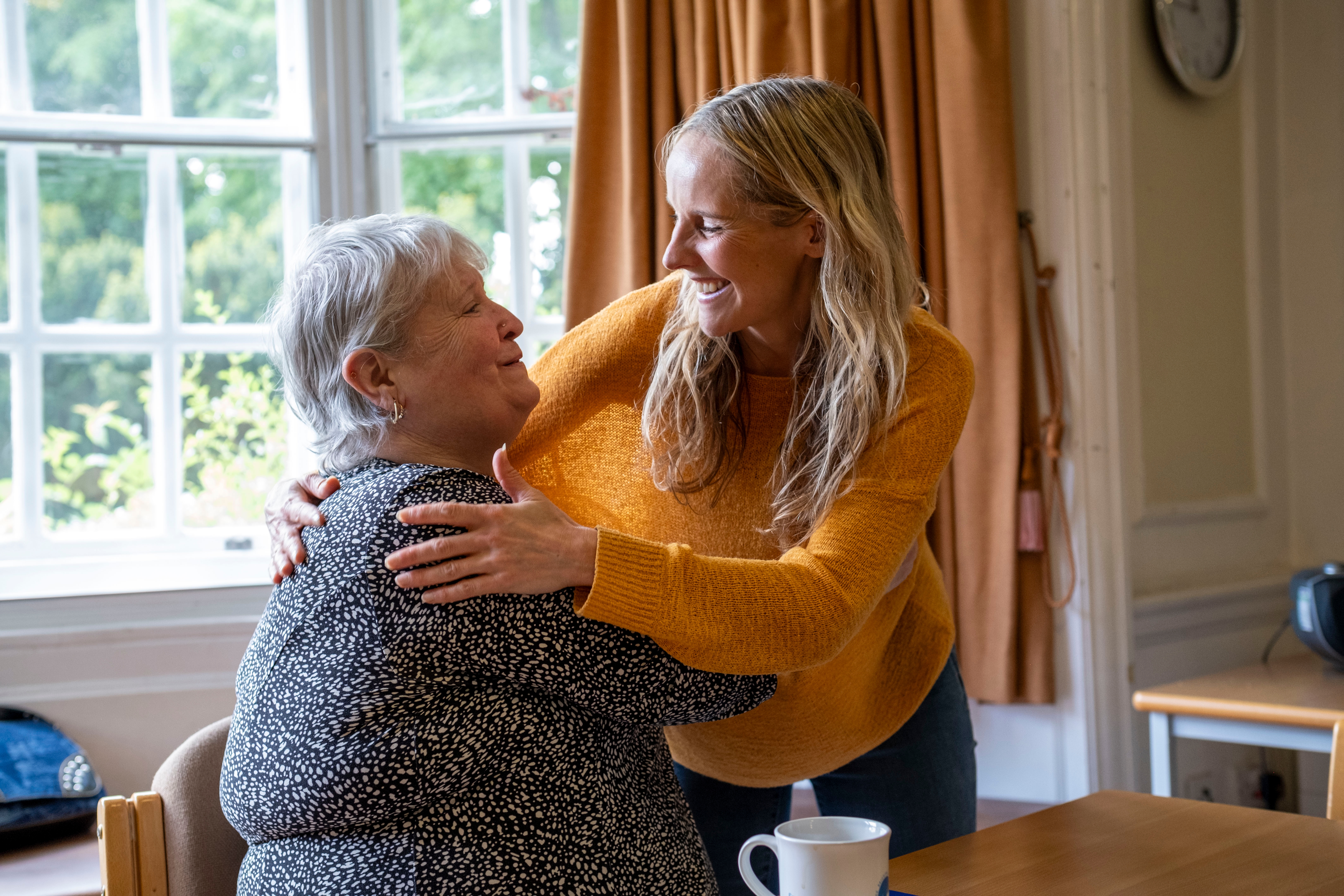 A young woman hugs an older woman at a table in a cozy room, sharing a warm moment
