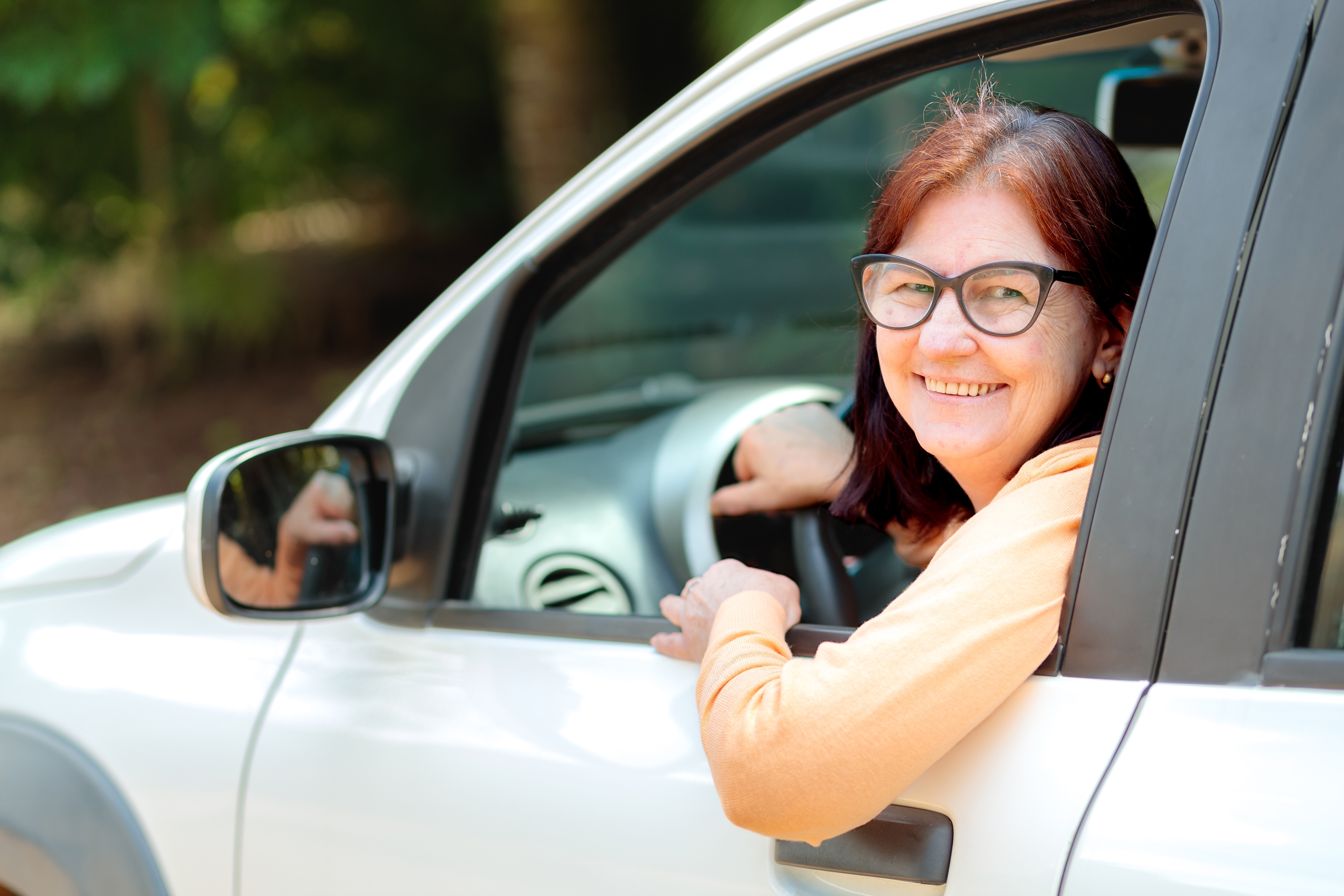 Smiling woman with glasses looks out from a car window, parked in a natural setting