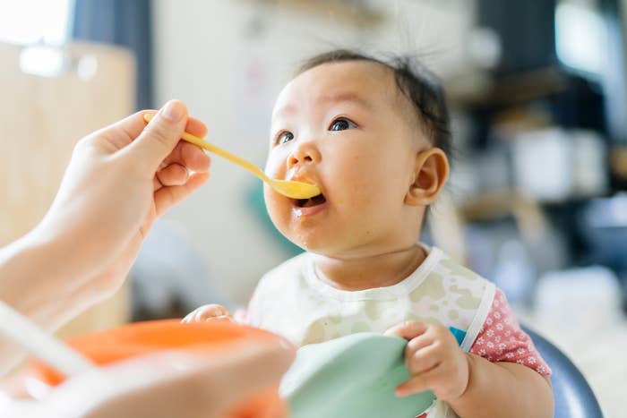Baby being fed by a spoon while sitting in a high chair, wearing a bib with a patterned design