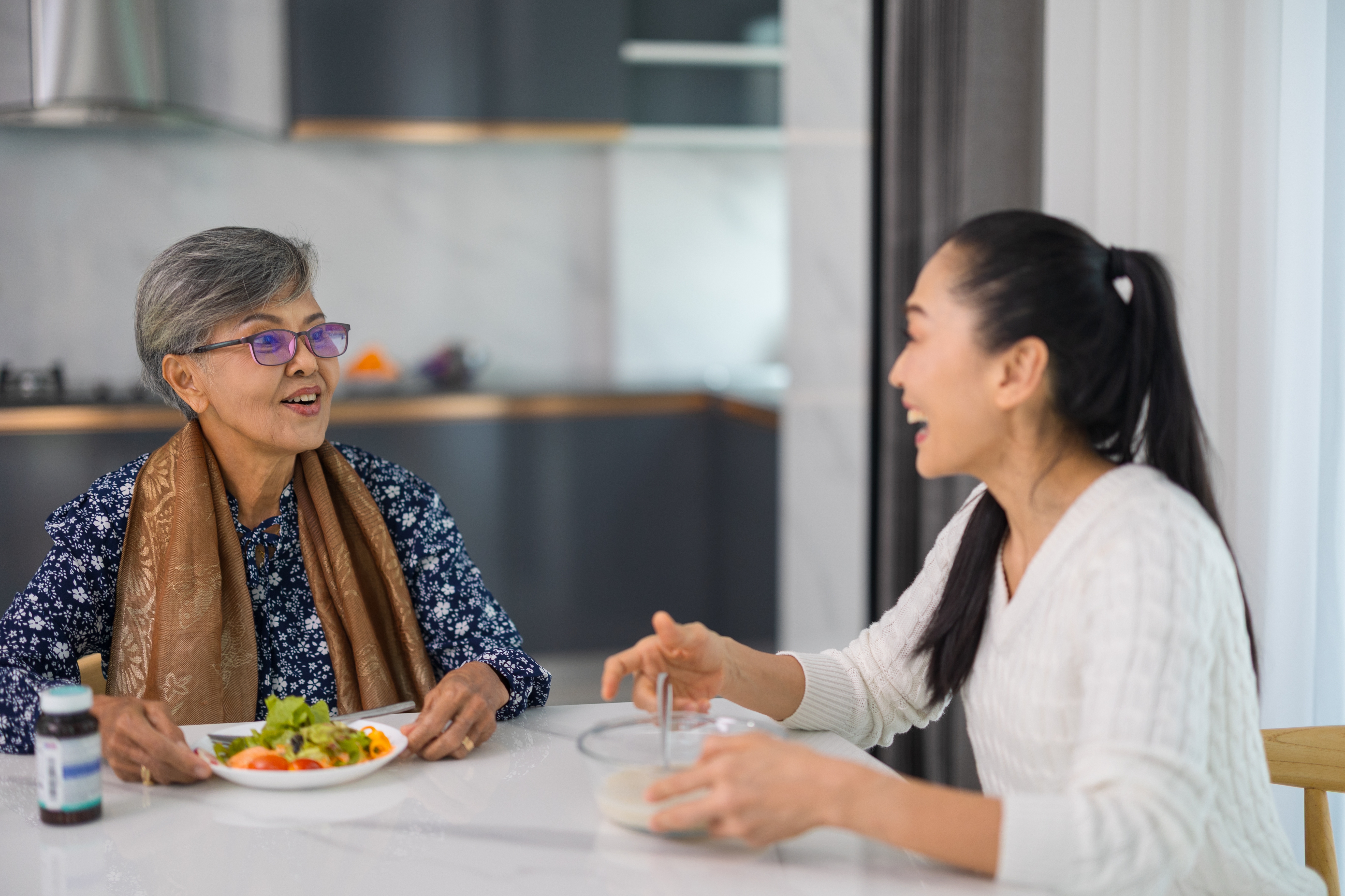 Two smiling women sit at a table, enjoying a meal together in a bright kitchen. One is holding a bowl, suggesting a friendly conversation