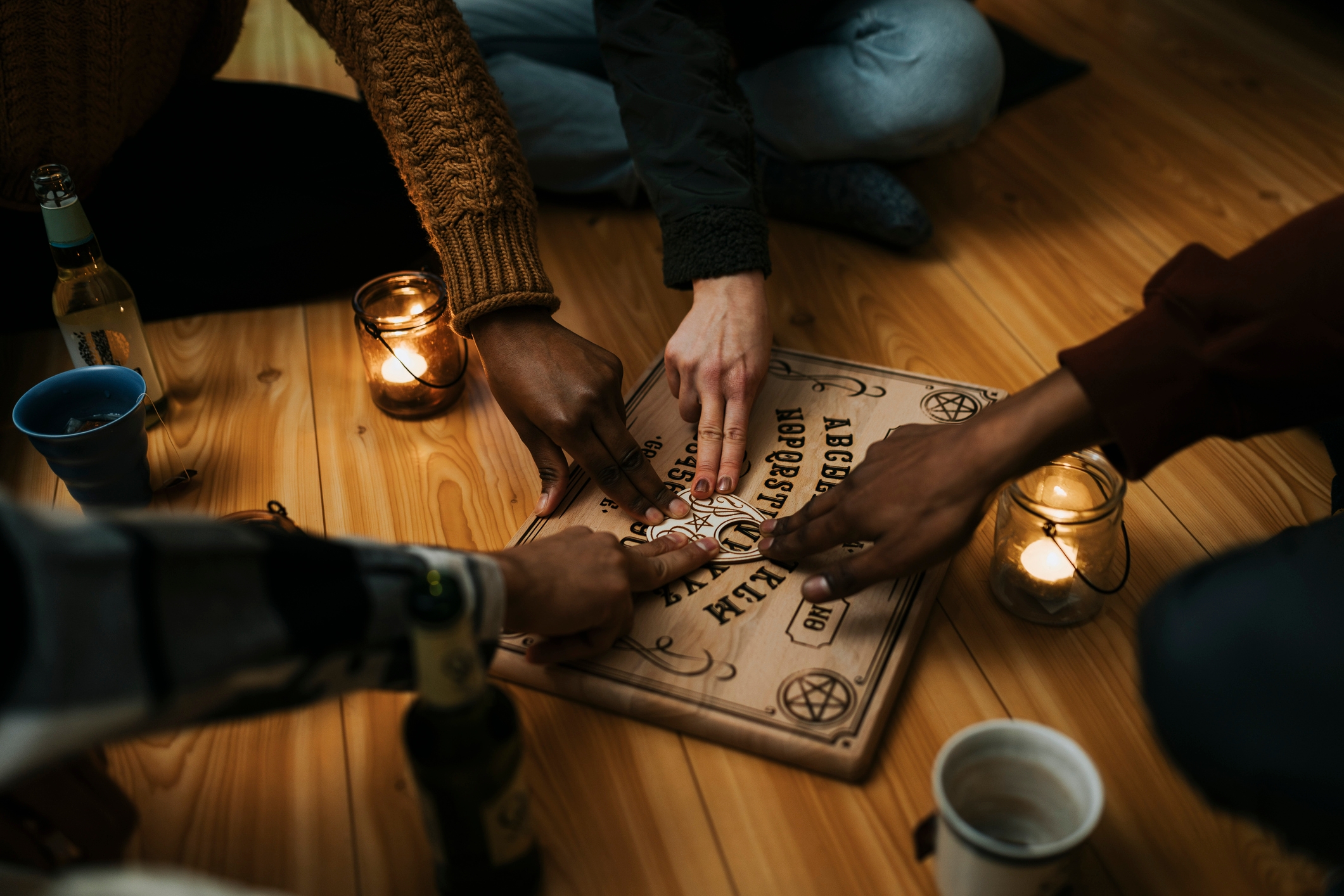 Hands of diverse individuals using a spirit board with lit candles nearby, creating a mystical atmosphere