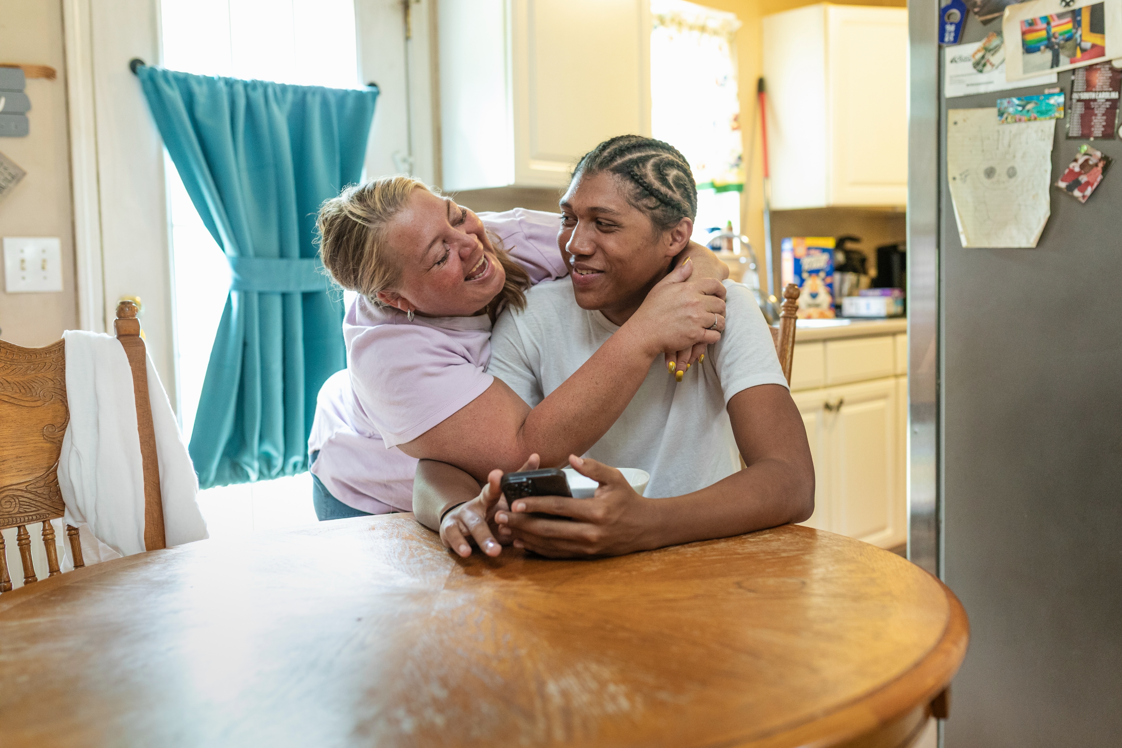 Two people smile warmly at each other in a kitchen. One stands, playfully embracing the other seated at a dining table, holding a phone