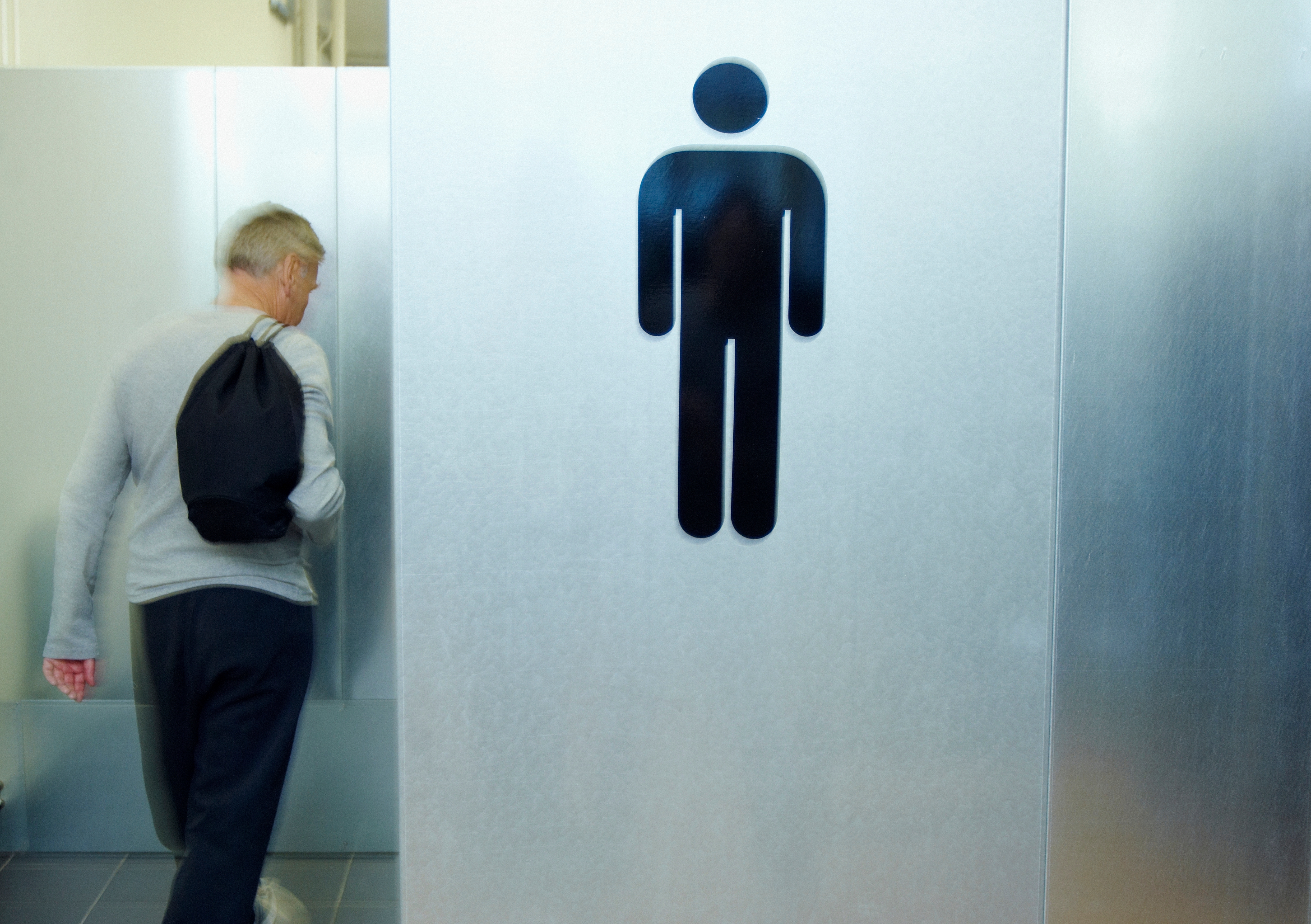 Person with backpack heading into a restroom with a large male figure sign on the door