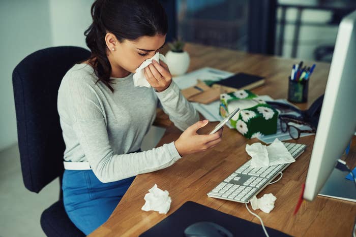 Person sitting at a desk, holding a phone, and blowing their nose into a tissue. Desk is cluttered with tissues, suggesting they have a cold