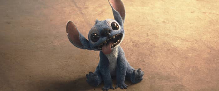 Animated character Stitch sitting with tongue out and playful expression on a sandy surface