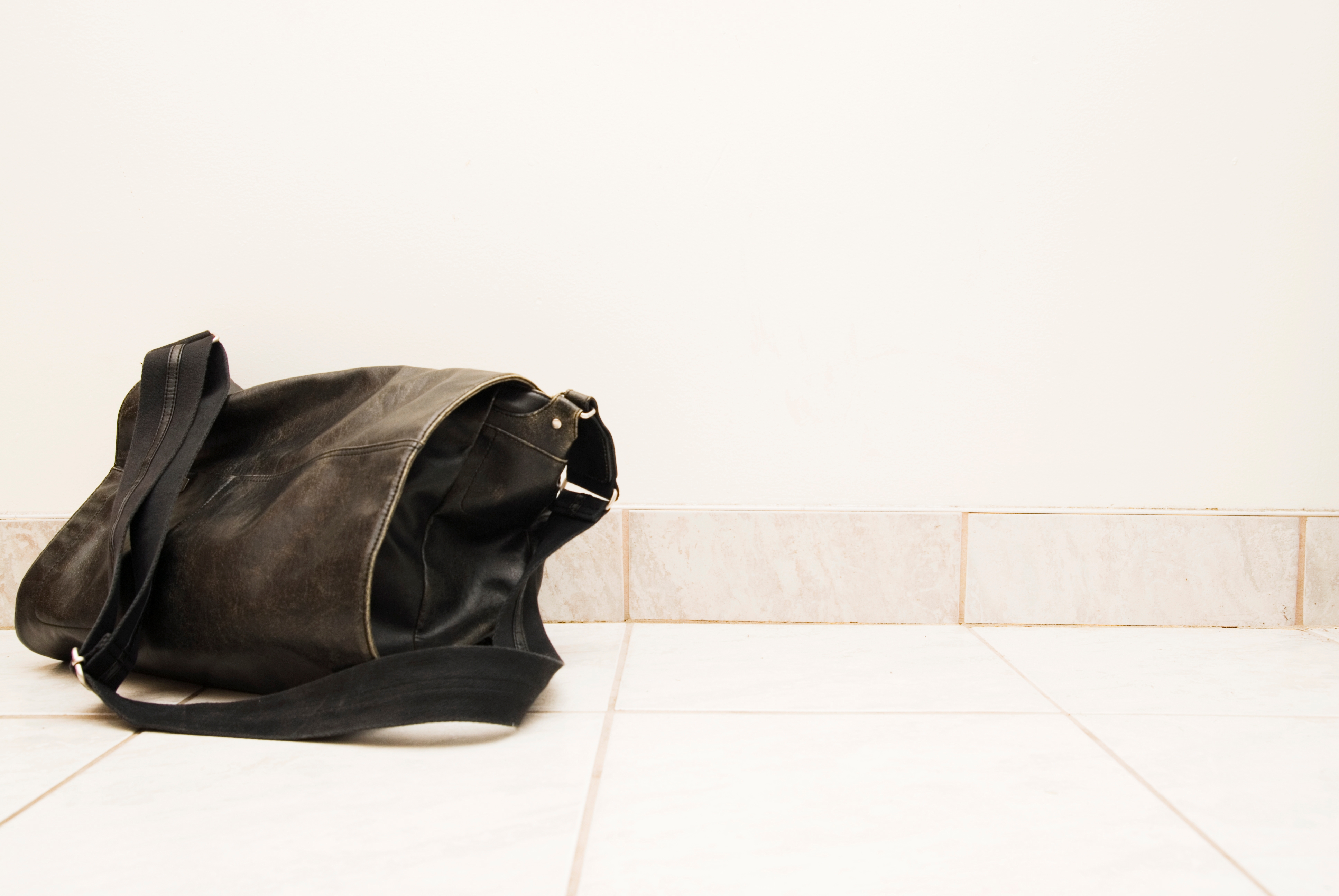 A black leather bag with a strap lies on a tiled floor against a plain wall