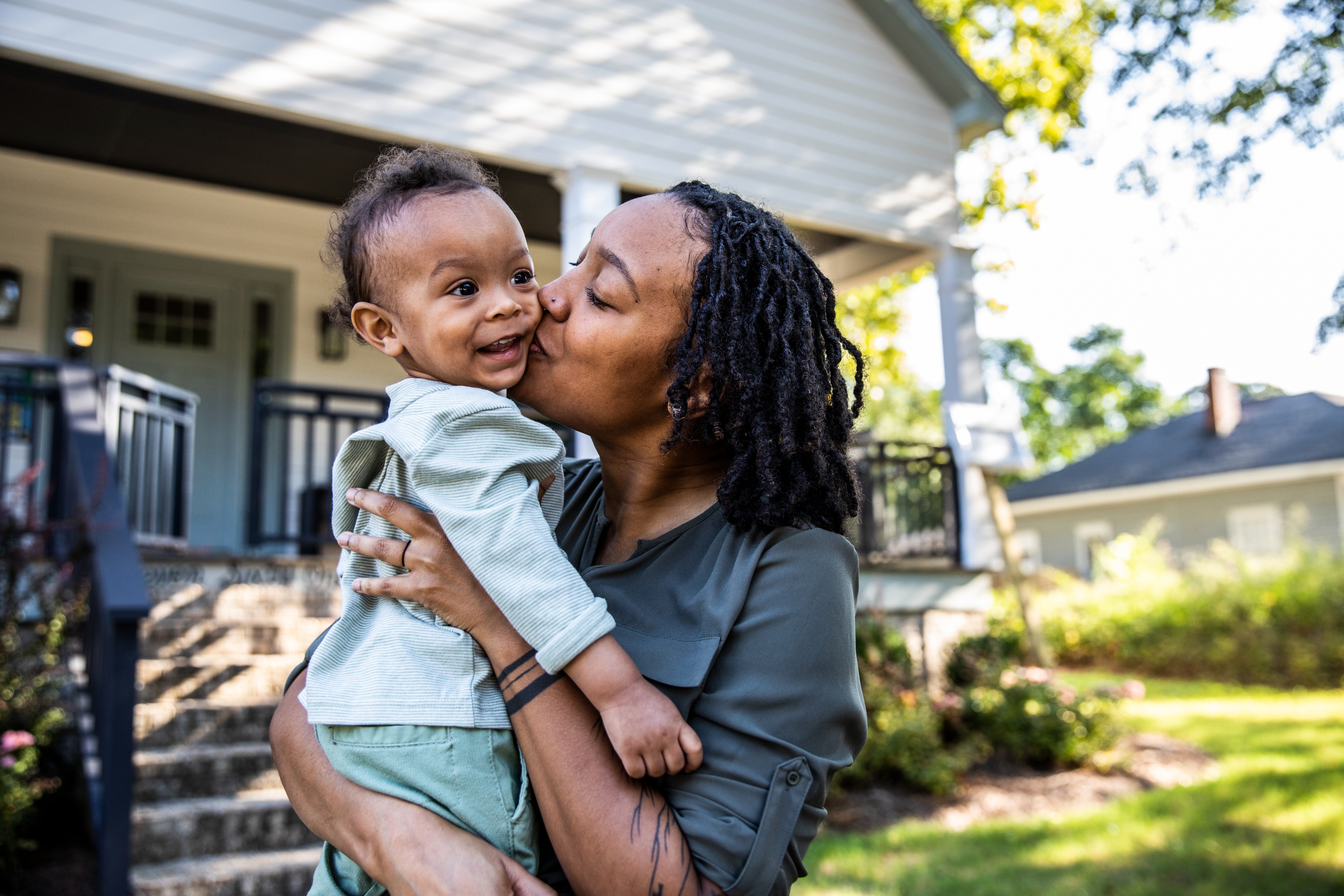 Parent affectionately kisses smiling toddler while holding them outside a house