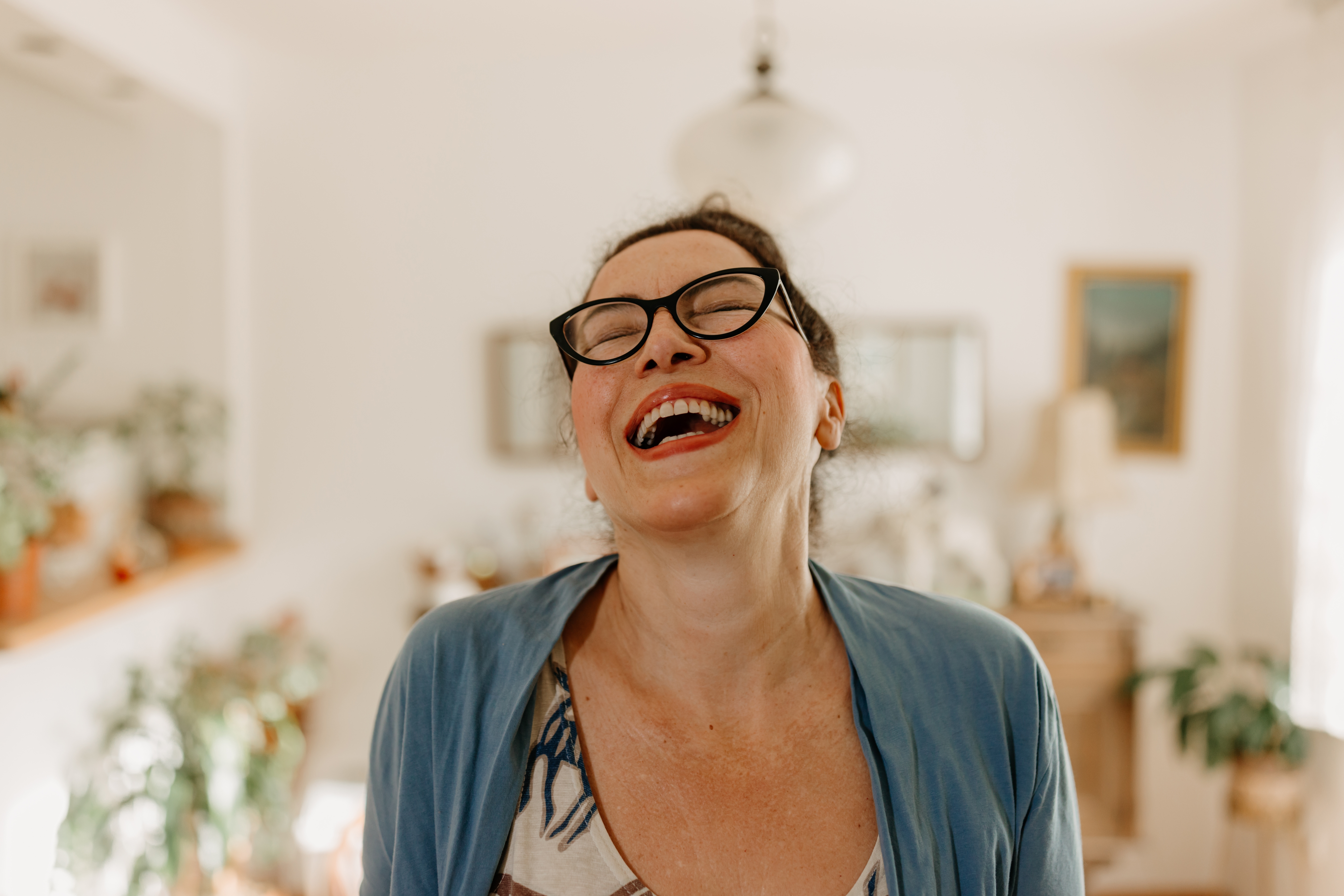 Person wearing glasses and a casual outfit, laughing in a sunny room with plants and framed pictures in the background