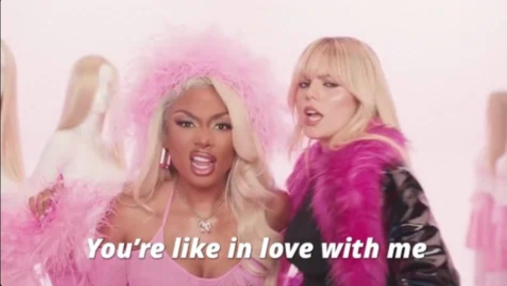 Two women with glamorous hairstyles pose confidently. One wears a feathered outfit, the other a shiny jacket. Text: "You’re like in love with me."