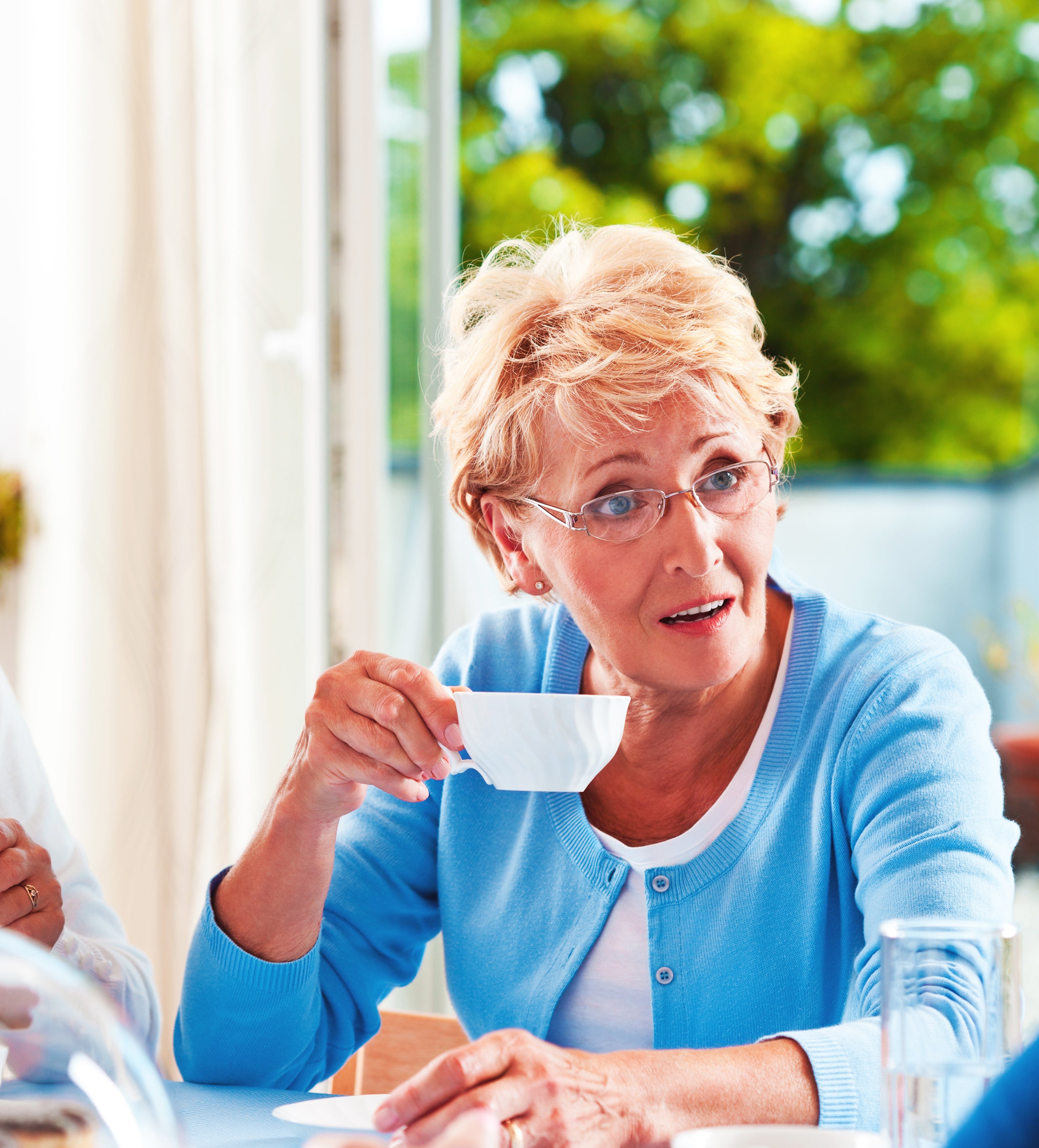 Woman with glasses holding a teacup, sitting at a table, engaged in conversation. She appears attentive and curious