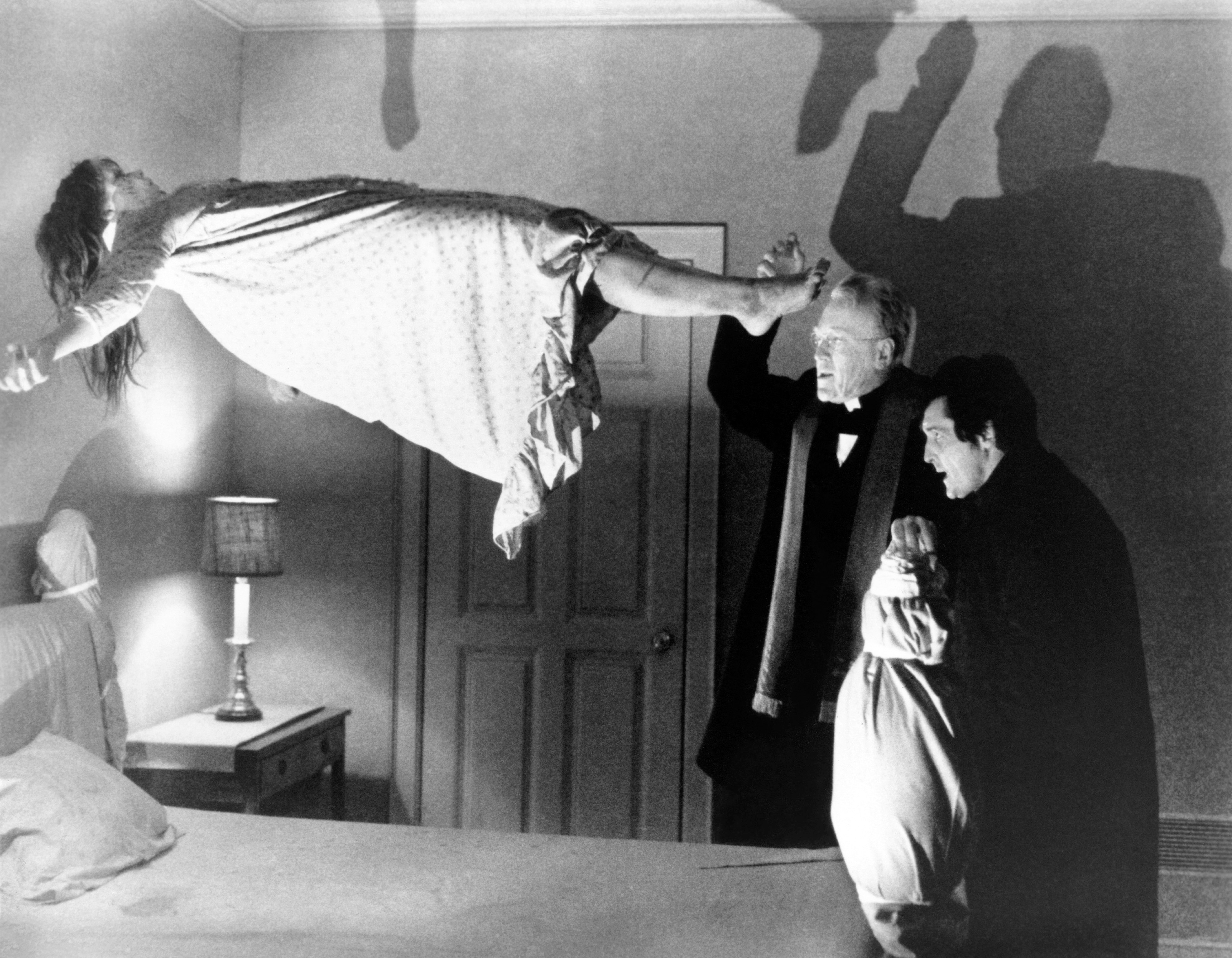 A girl levitates above a bed in a dramatic scene, with two concerned men observing, one in a robe and another holding a small book