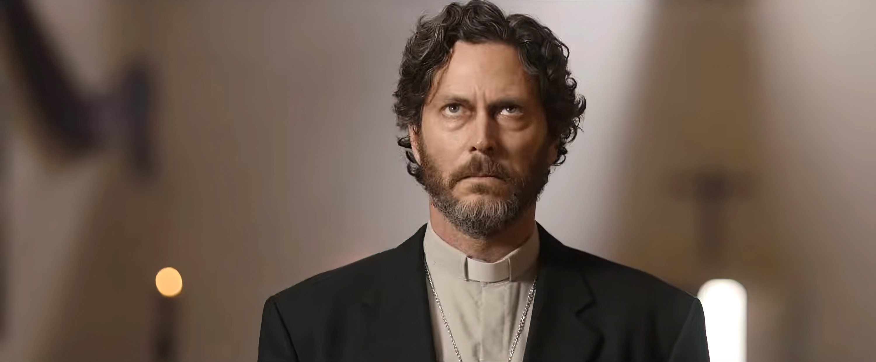 A solemn man in priest attire stands in a church, looking upward with a serious expression