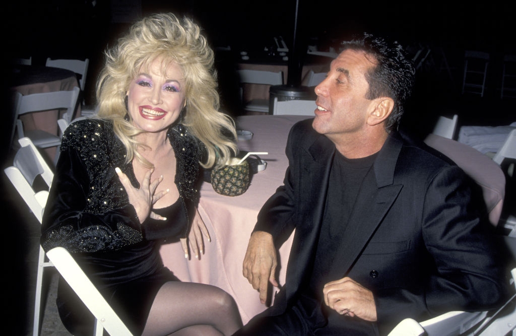 Two people seated at a table, one with voluminous hair and a sparkling dress, the other in a dark suit, both smiling and engaged in conversation