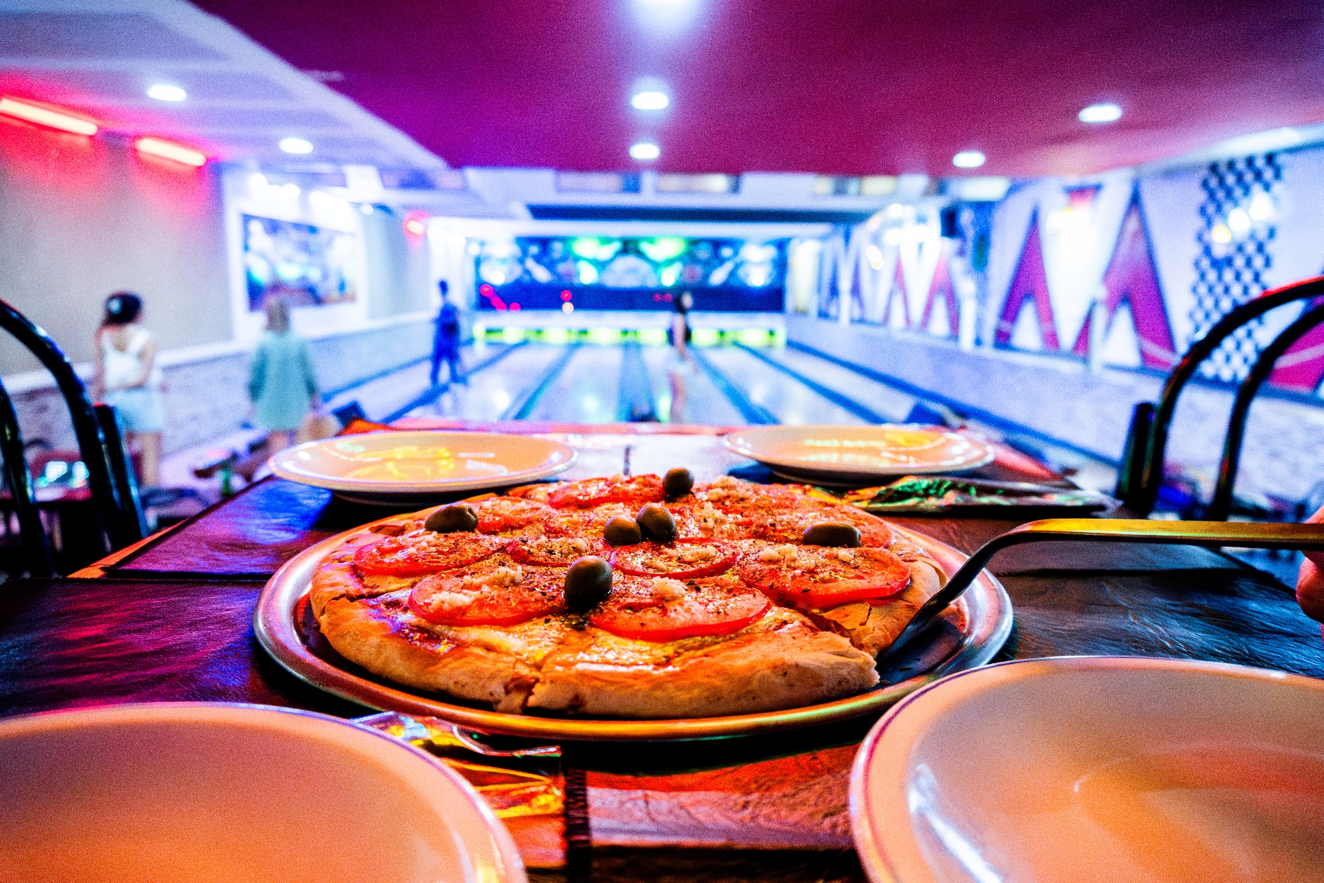 A pizza on a table in a bowling alley, with bowling lanes and people in the background
