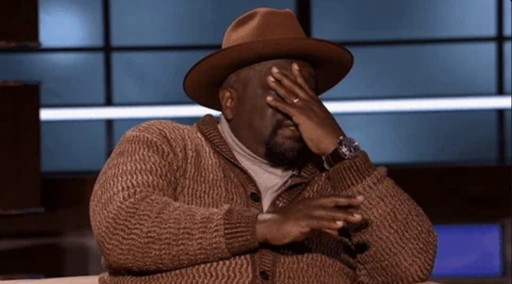 A person in a sweater and hat sits, covering their face with one hand, possibly expressing laughter or disbelief