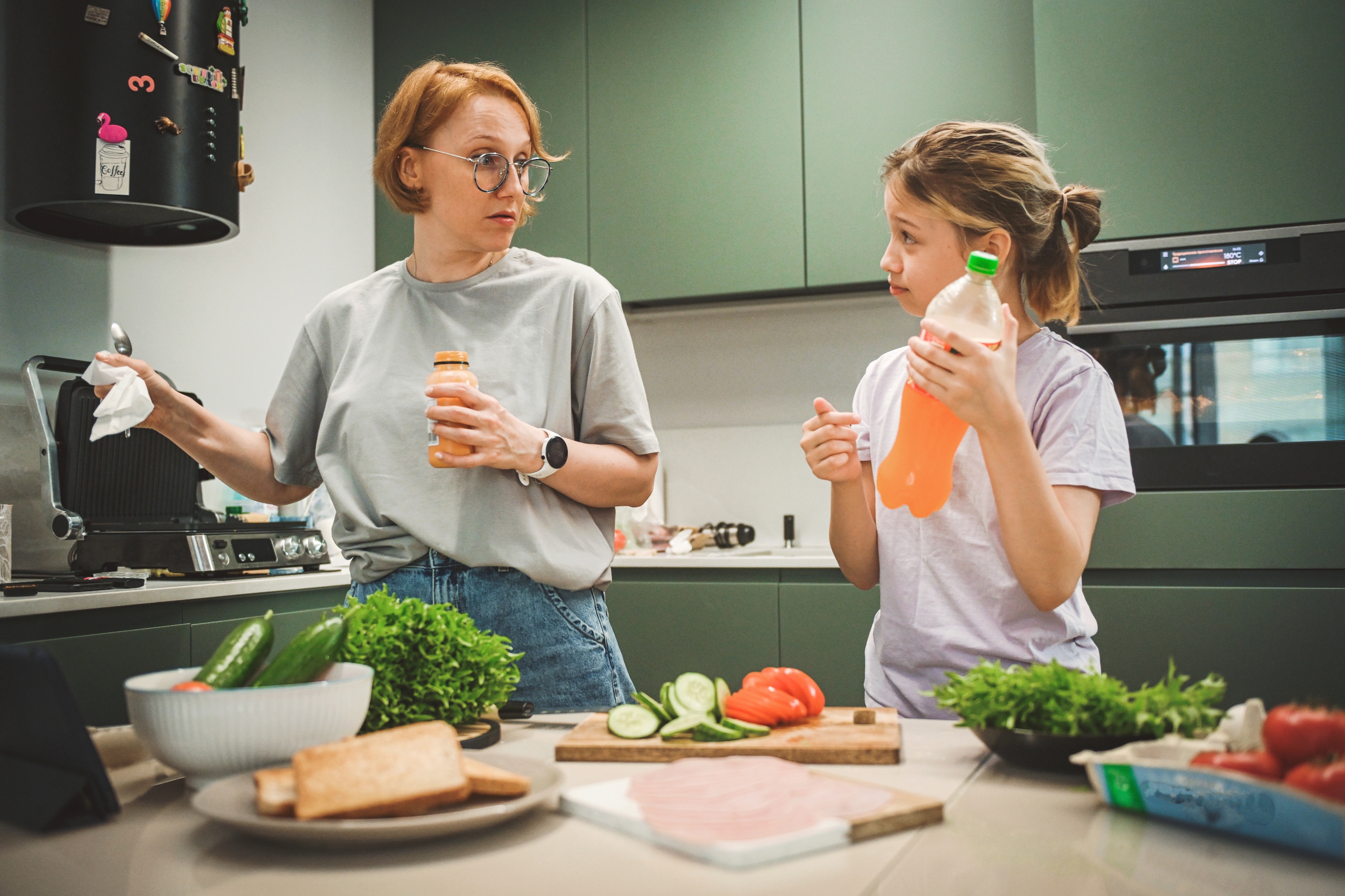 Woman and girl in a kitchen preparing a meal, surrounded by ingredients like cucumbers, lettuce, tomatoes, and bread. They appear to be in conversation