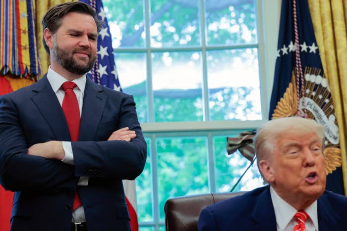 JD Vance with arms crossed, looking down with a smile at Donald Trump, who is seated in the Oval Office and speaking