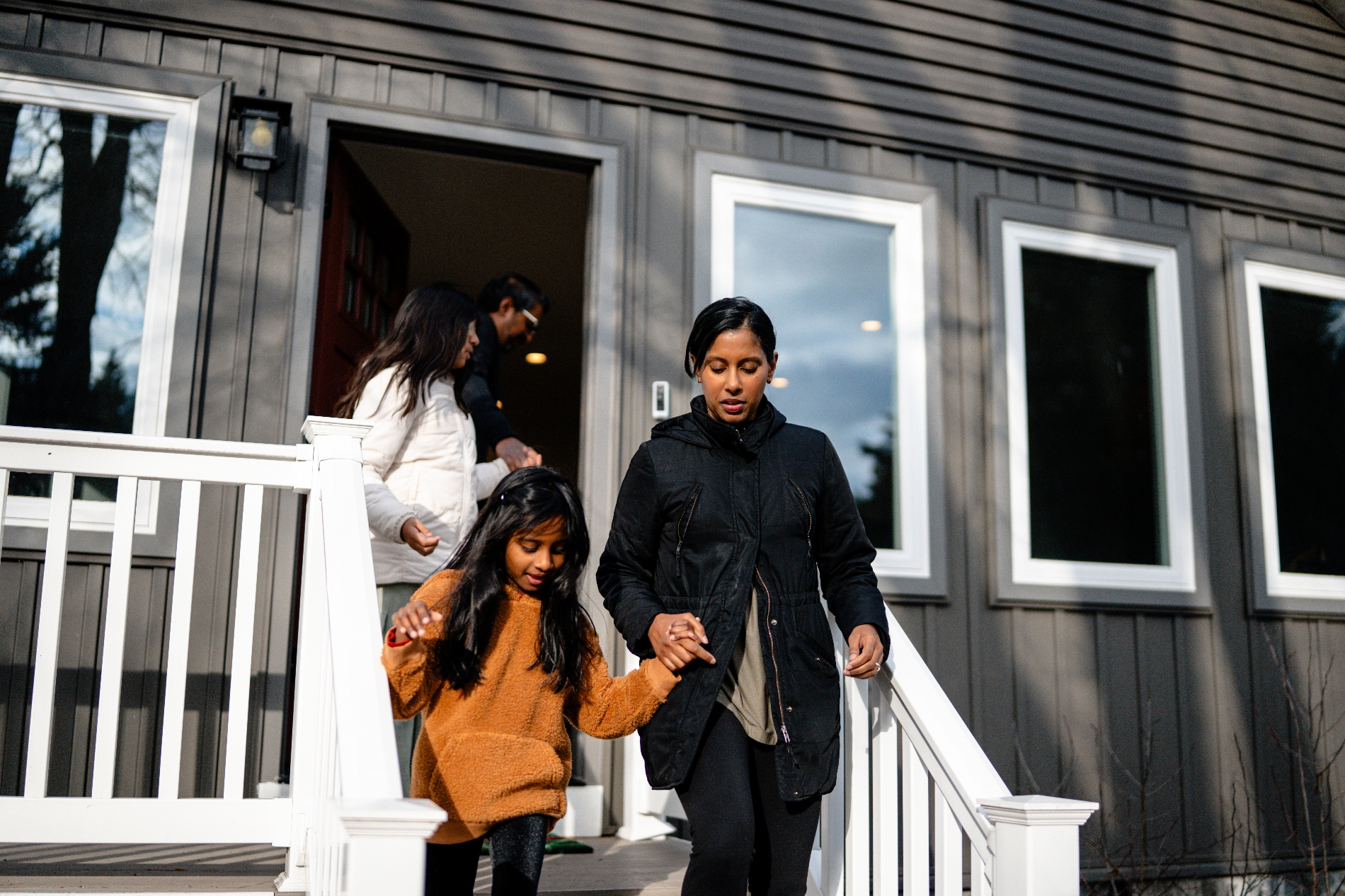 A family of four walks down the steps of a house. A woman holds a child's hand. Everyone is wearing casual, warm clothing