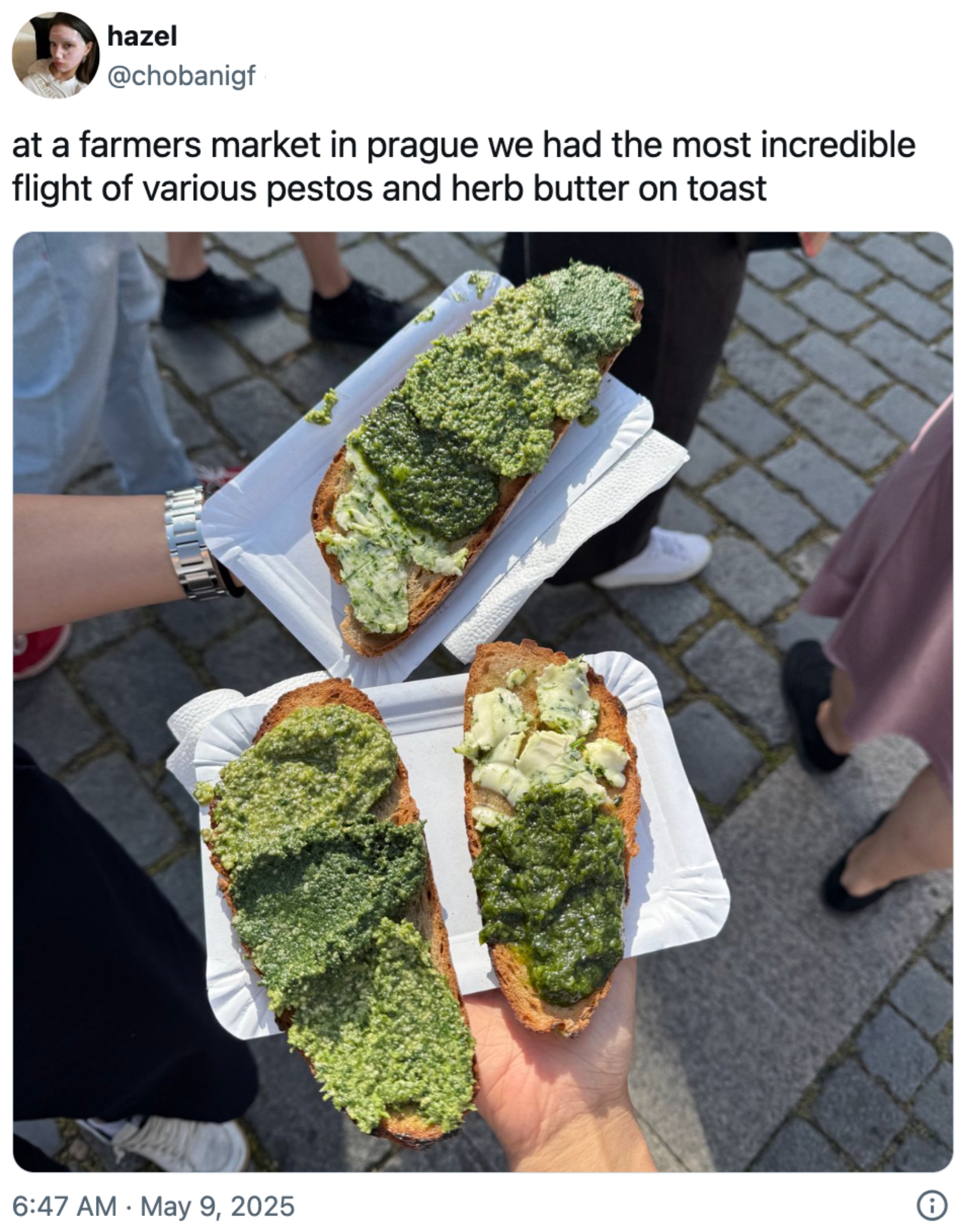 Three slices of toast topped with various pestos and herb butter, held by people at a farmers market in Prague