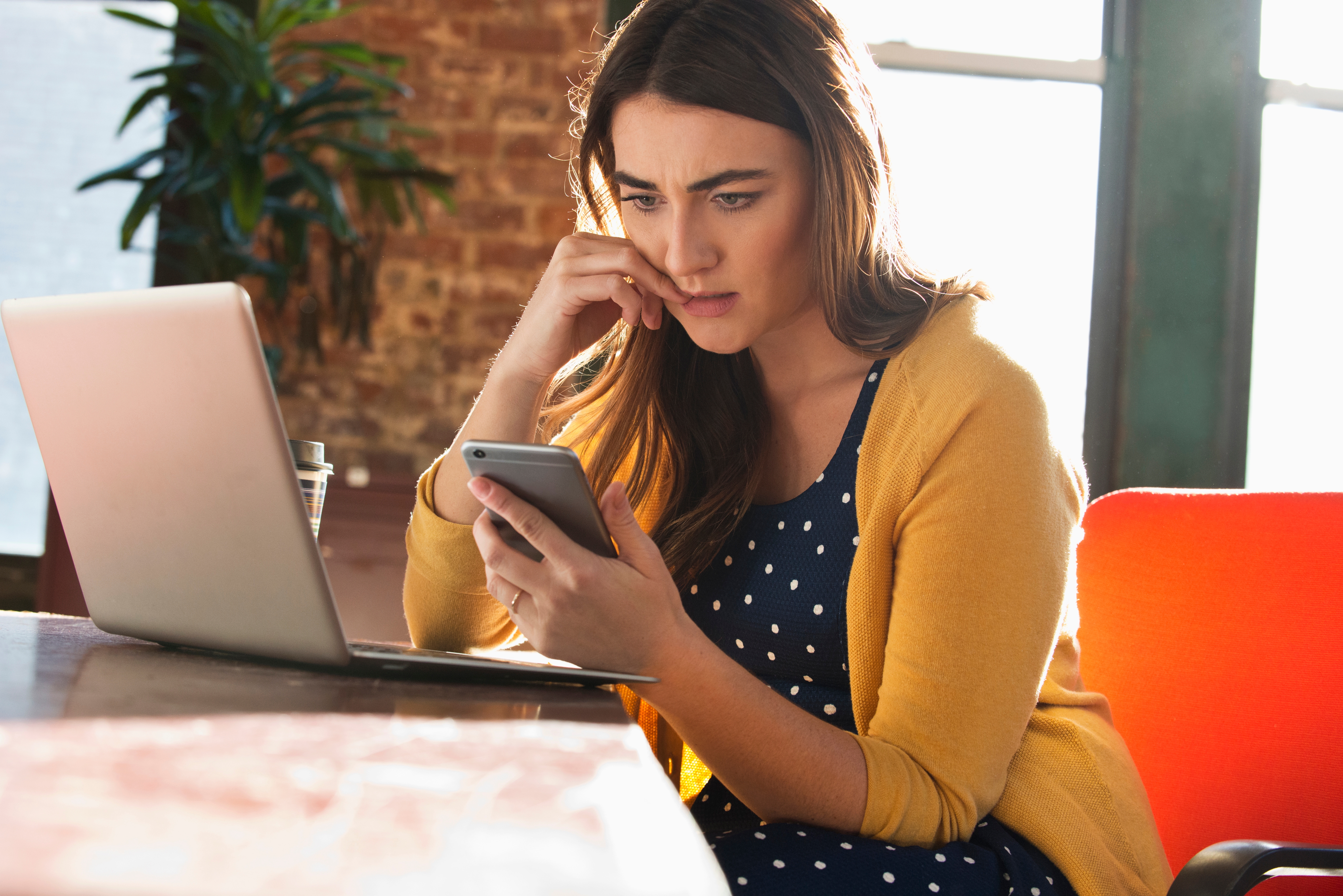 Woman in a polka dot dress and cardigan looks concerned while checking her phone at a table with a laptop