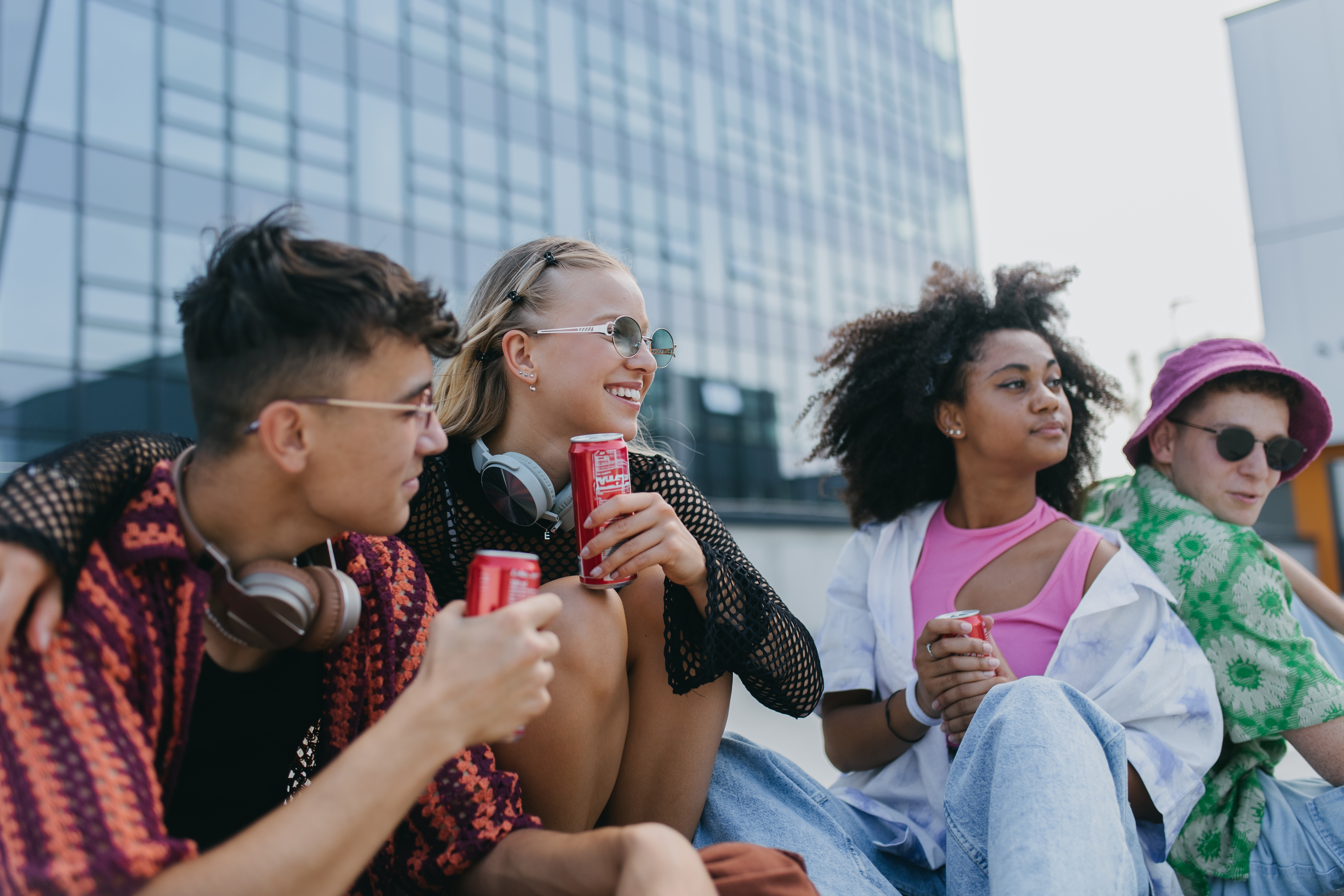 Four people sit together outdoors, each holding a drink can. They're relaxed and stylish, wearing casual summer clothing