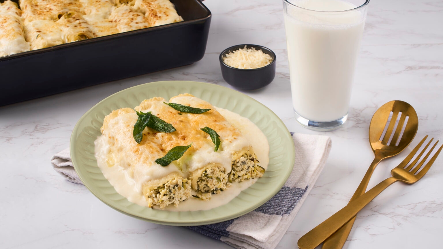 Plate of creamy stuffed pasta with herbs, next to a baking dish, a small bowl of cheese, a glass of milk, and golden cutlery on a napkin