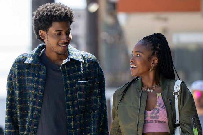 Two people walking and smiling at each other. The person on the left wears a checkered jacket, and the person on the right wears a cropped top and jacket