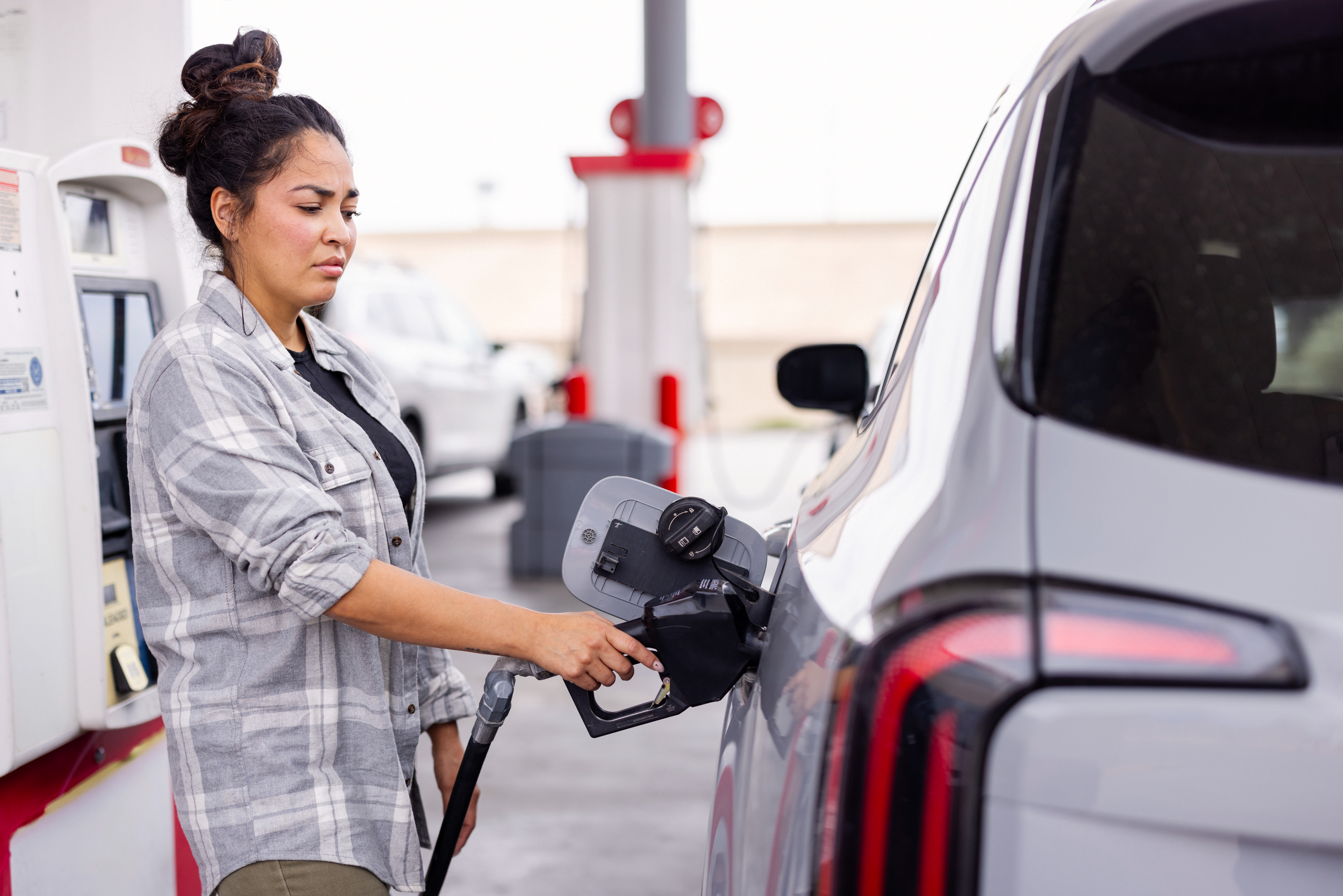 A person pumps gas into a car at a fuel station, appearing focused. They are wearing a plaid shirt and have their hair in a bun