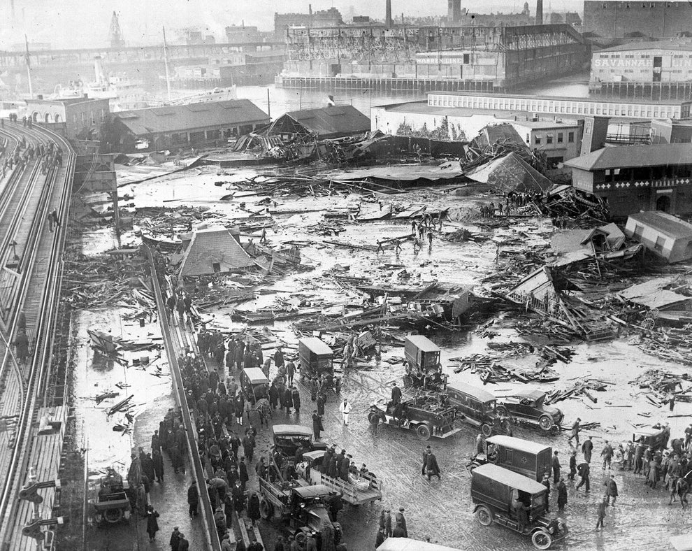 Crowd and vehicles gather at a waterfront disaster site with debris and collapsed structures spread across the area
