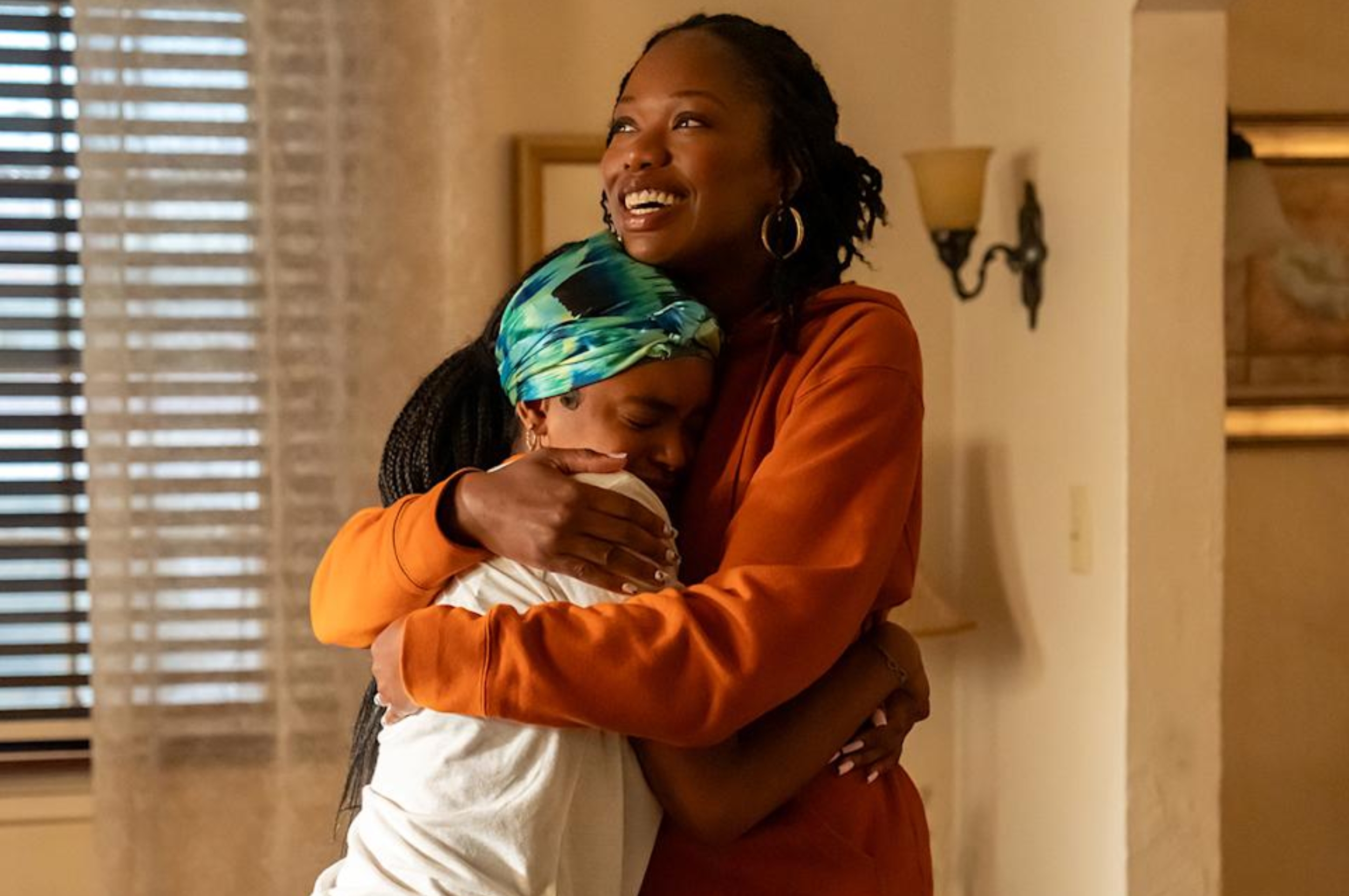 Two people embrace warmly in a cozy room, one smiling joyfully while the other rests their head on their shoulder