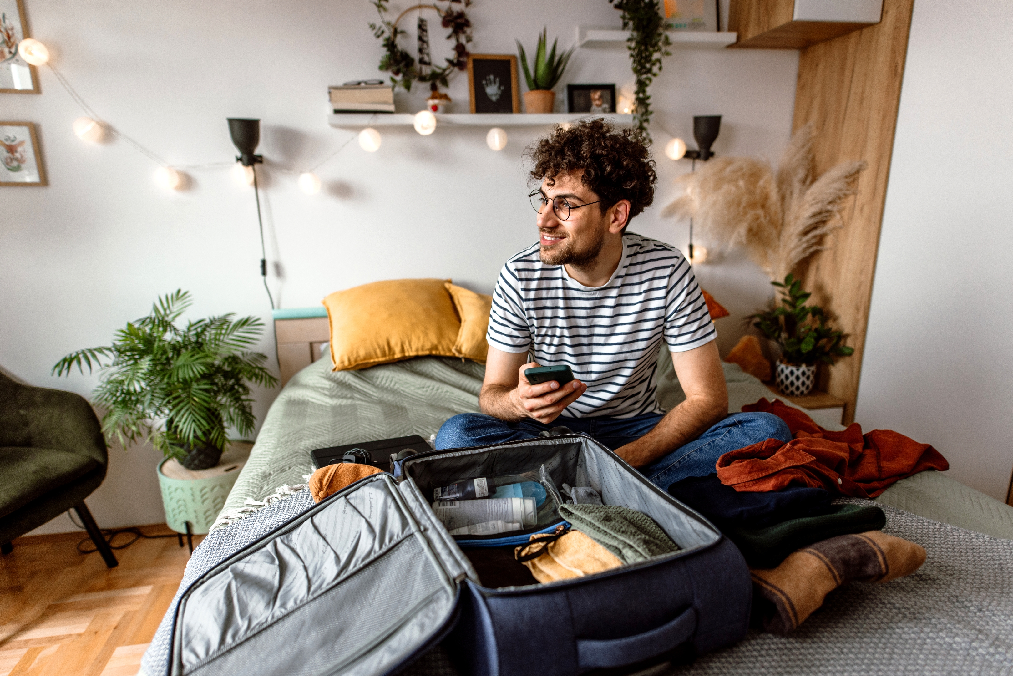 A person sits on a bed with an open suitcase, holding a phone, surrounded by plants and string lights in a cozy room