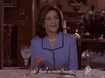 Woman in elegant attire sits at dining table, smiling and holding a fork while saying, "Oh! that's very funny."