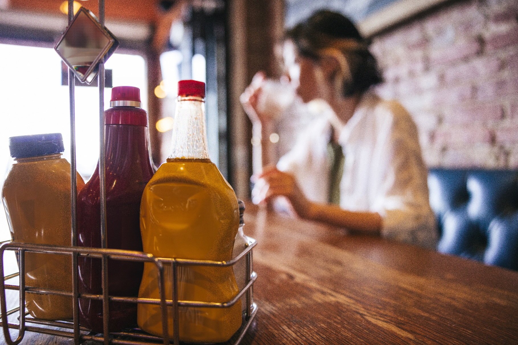 Ketchup and mustard bottles on a table in focus, with a person drinking in the blurred background at a restaurant