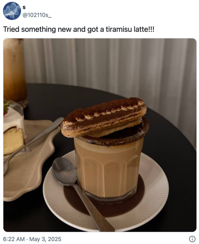 A latte topped with a tiramisu cookie on a saucer with a spoon, accompanied by a slice of cake in the background