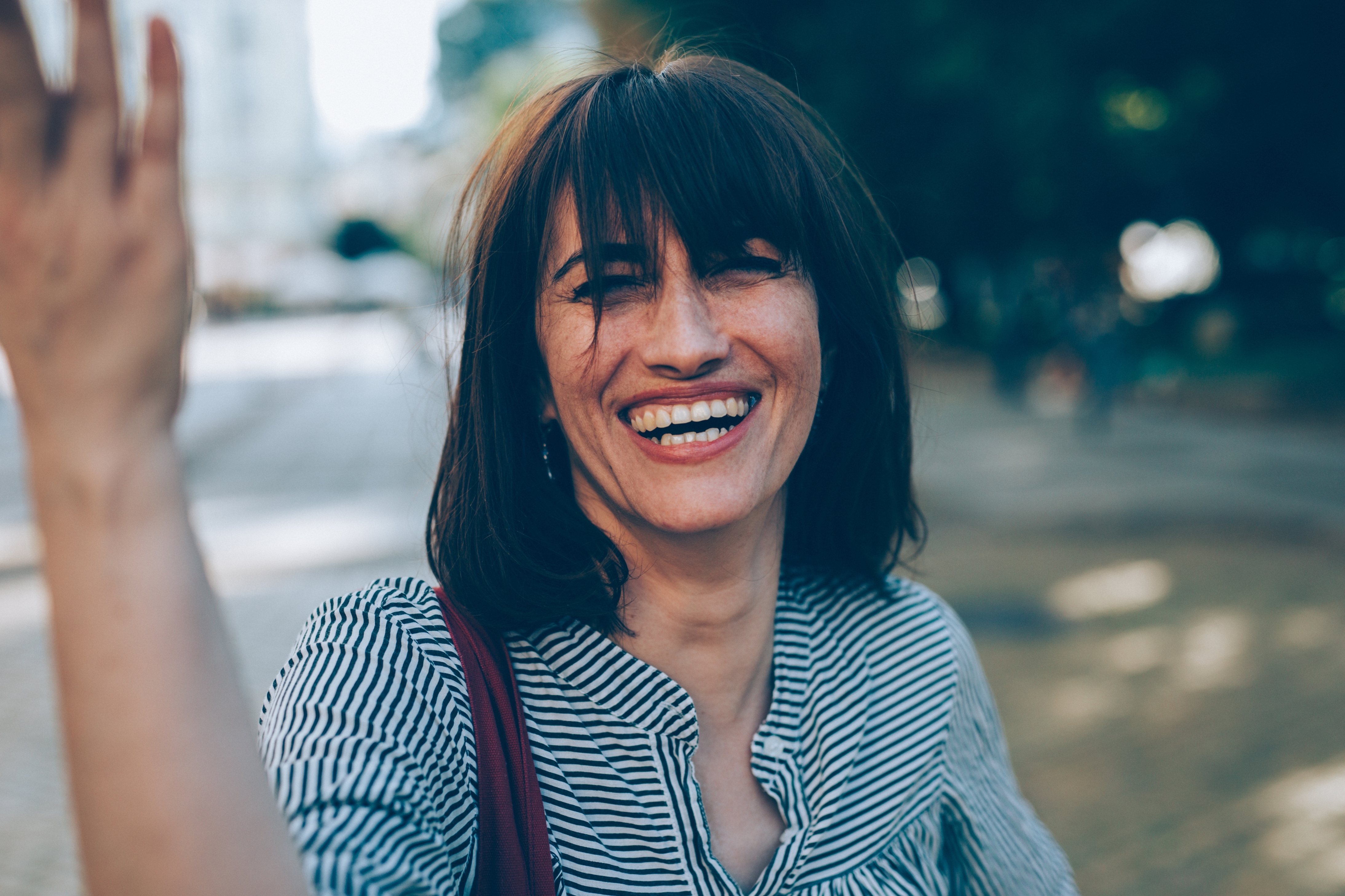 Woman smiling outside, wearing a striped top, with blurred trees and pavement in the background