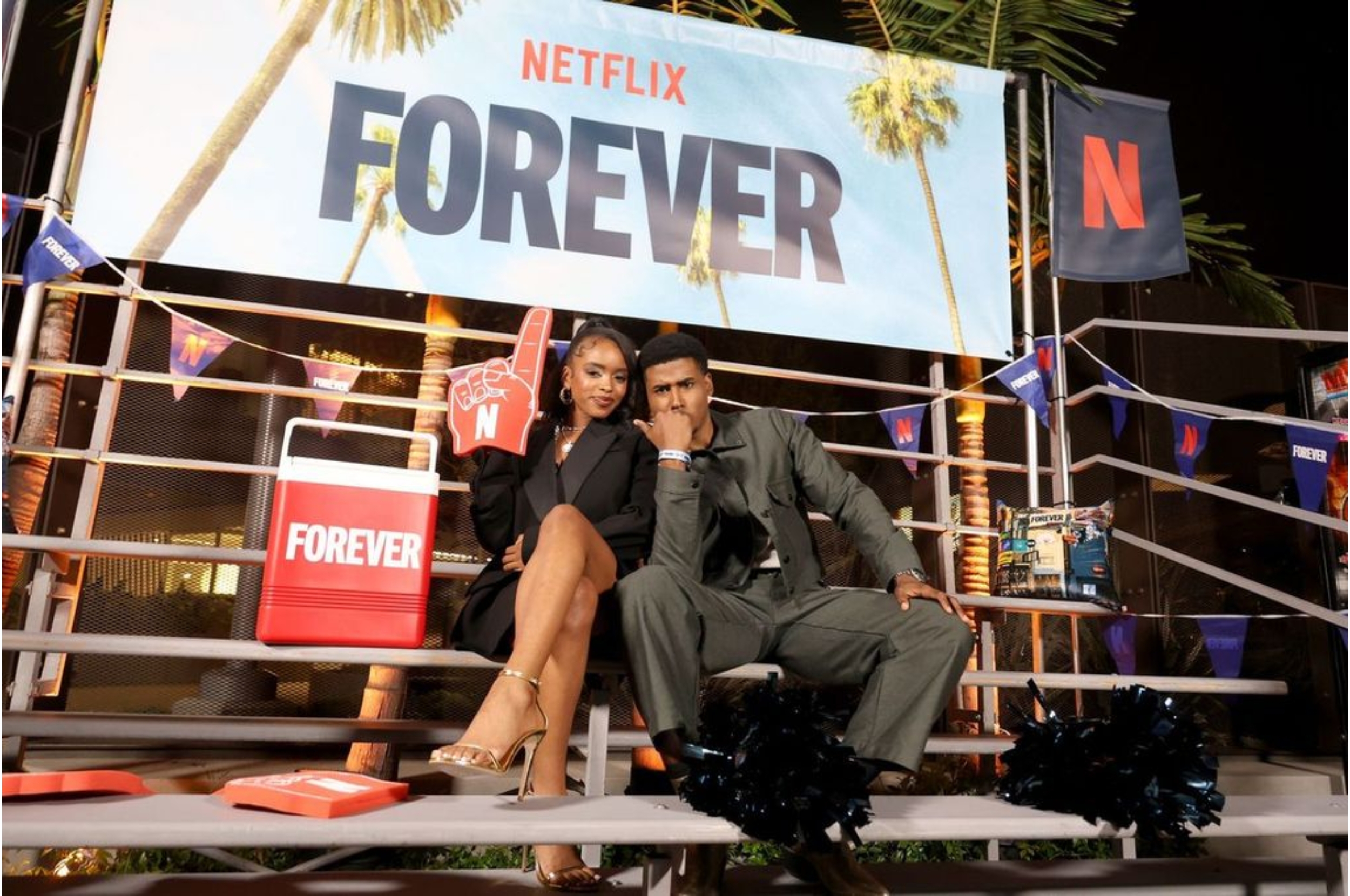 Two people sit on bleachers at a Netflix event, with one holding a novelty "N" sign. The banner reads "FOREVER."