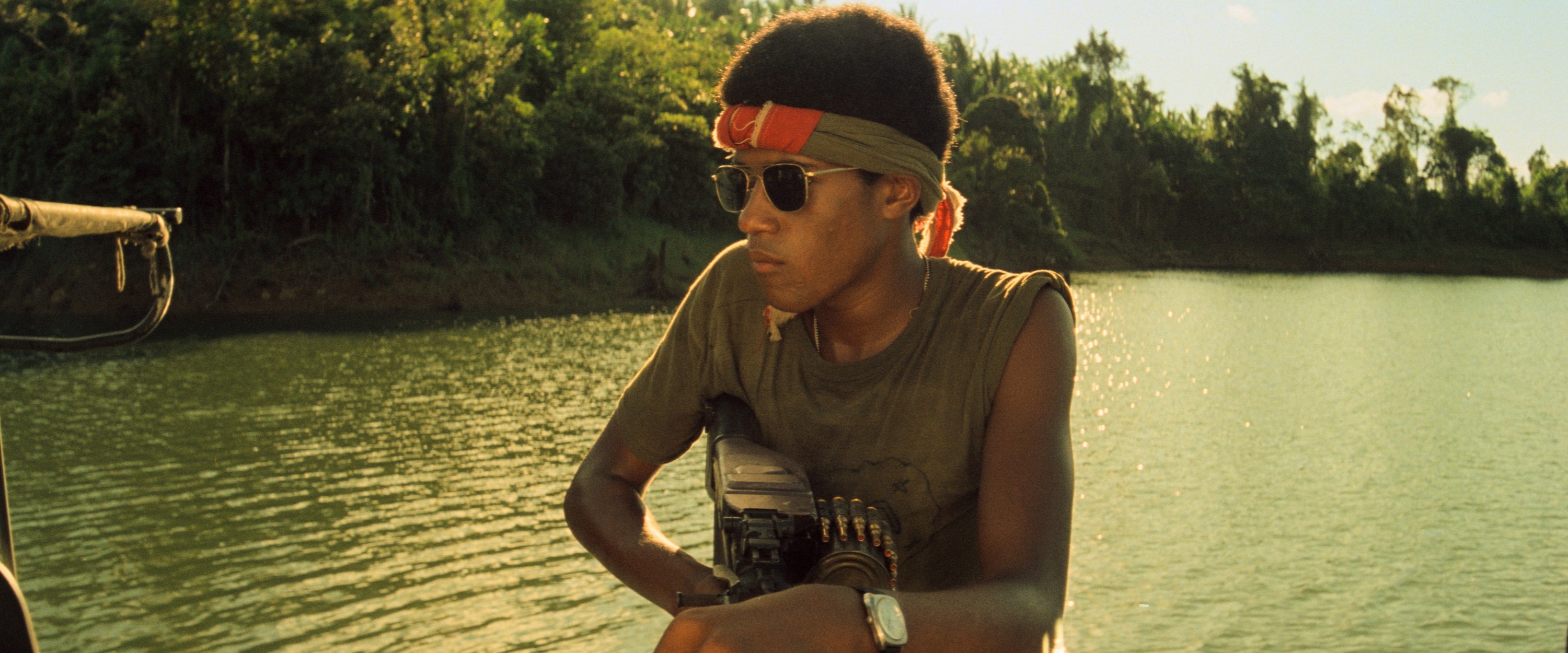 A person with sunglasses and a headband holds a weapon, looking out over a calm river, surrounded by trees