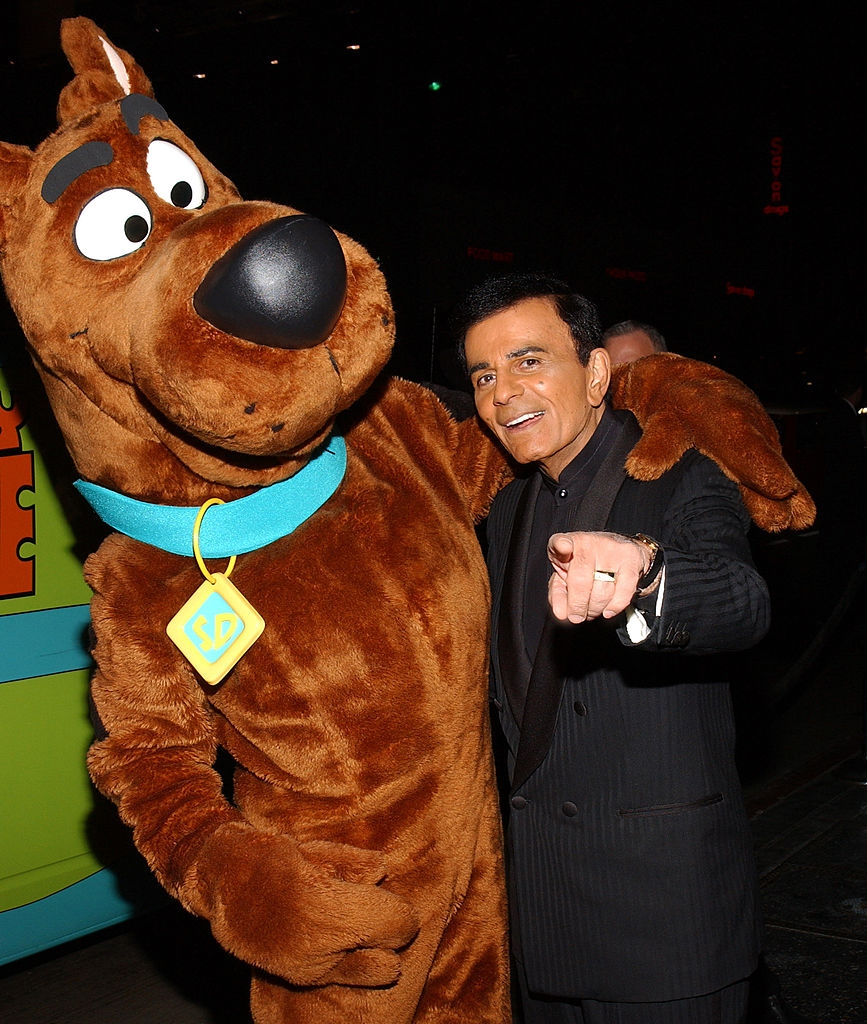 Person poses with a life-sized Scooby-Doo character, smiling and pointing at the camera. They're in a dimly lit, possibly outdoor setting